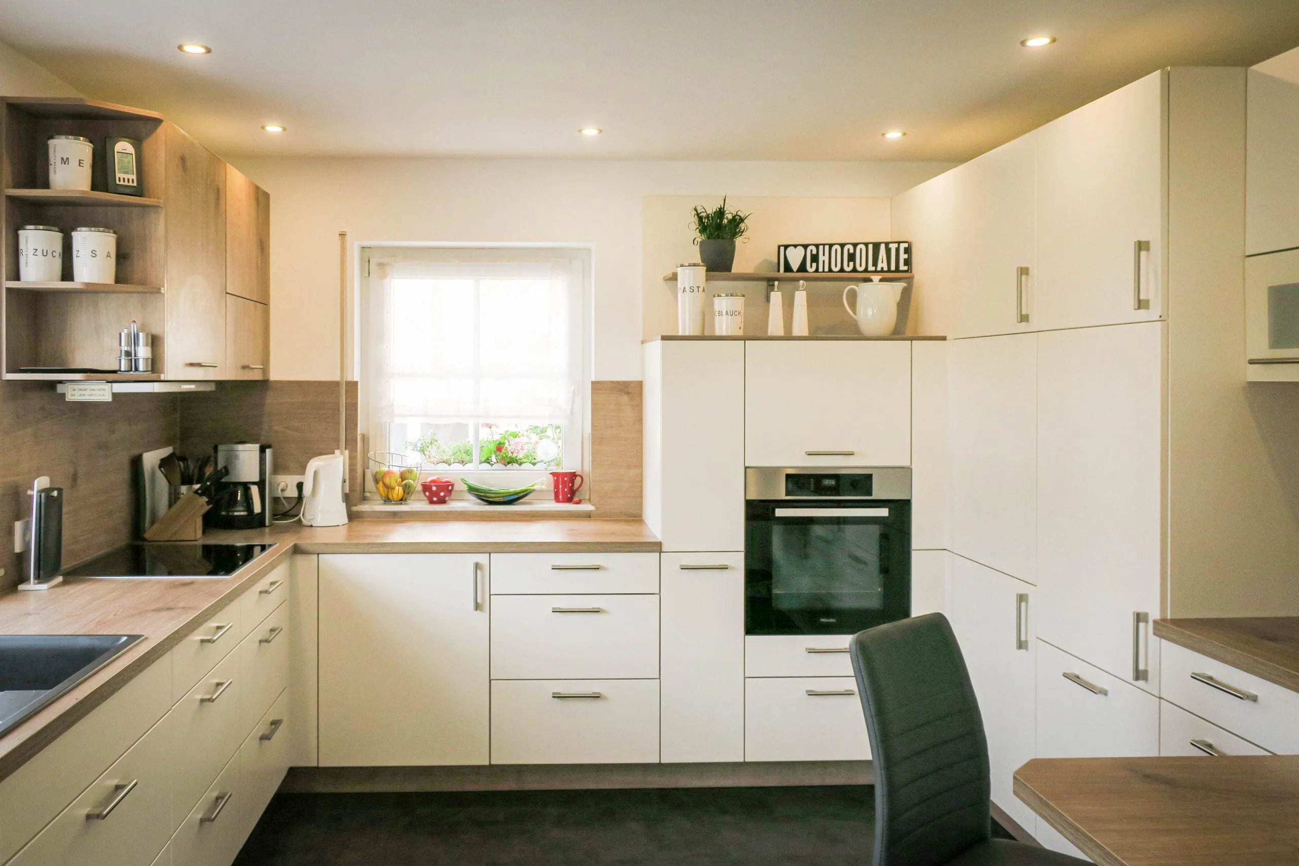 Gesamtansicht der weißen L-Küche mit Spülbereich am Fenster, Eckschrank mit Tellerbord, Backofen-Hochschrank, Holz-Bartheke und integriertem Esstisch