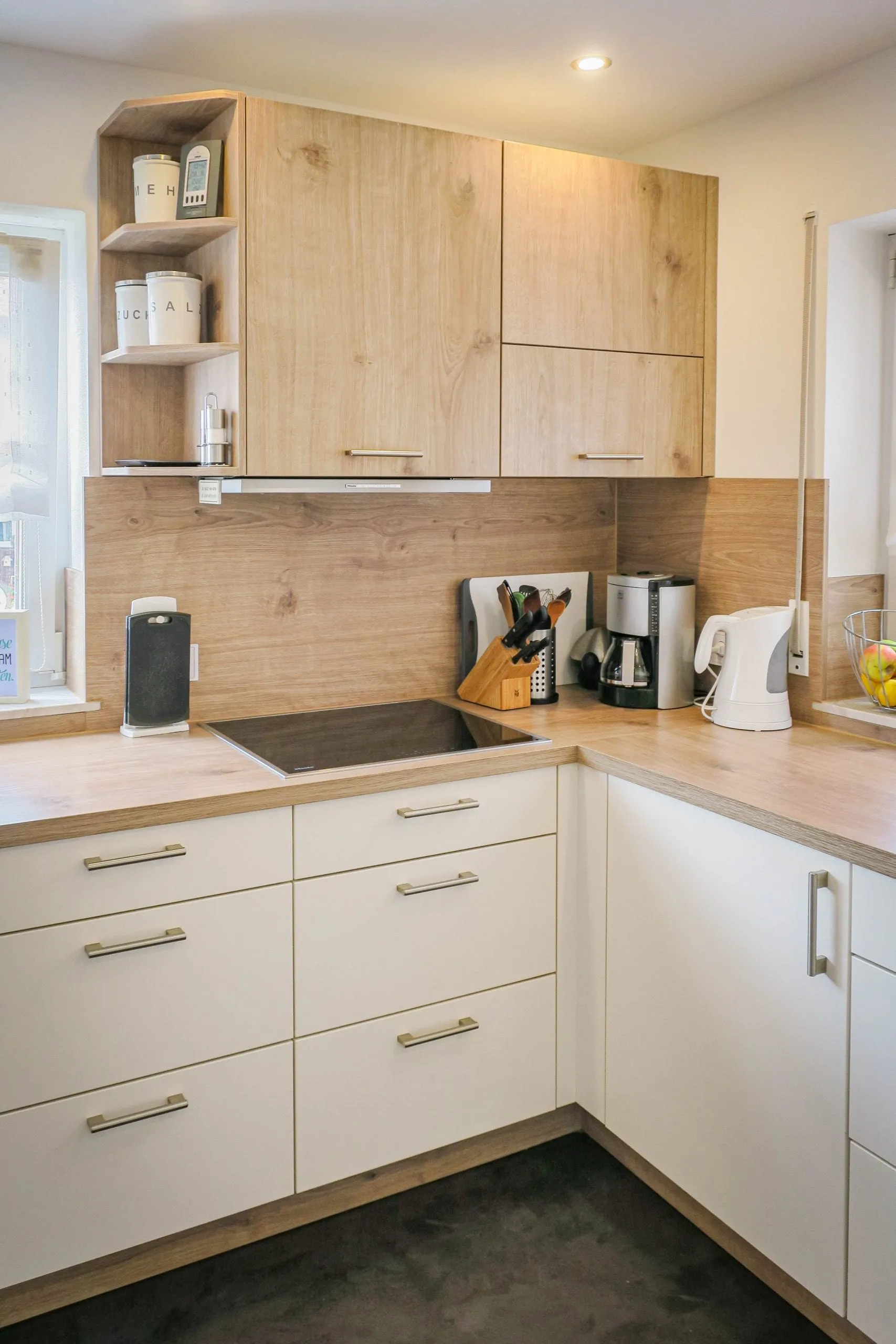 Eck-Kochbereich mit Induktionskochfeld auf Holz-Arbeitsplatte und Holz-Rückwand, Holz-Hängeschrank mit offenem Eck-Tellerbord, weiße Unterschränke