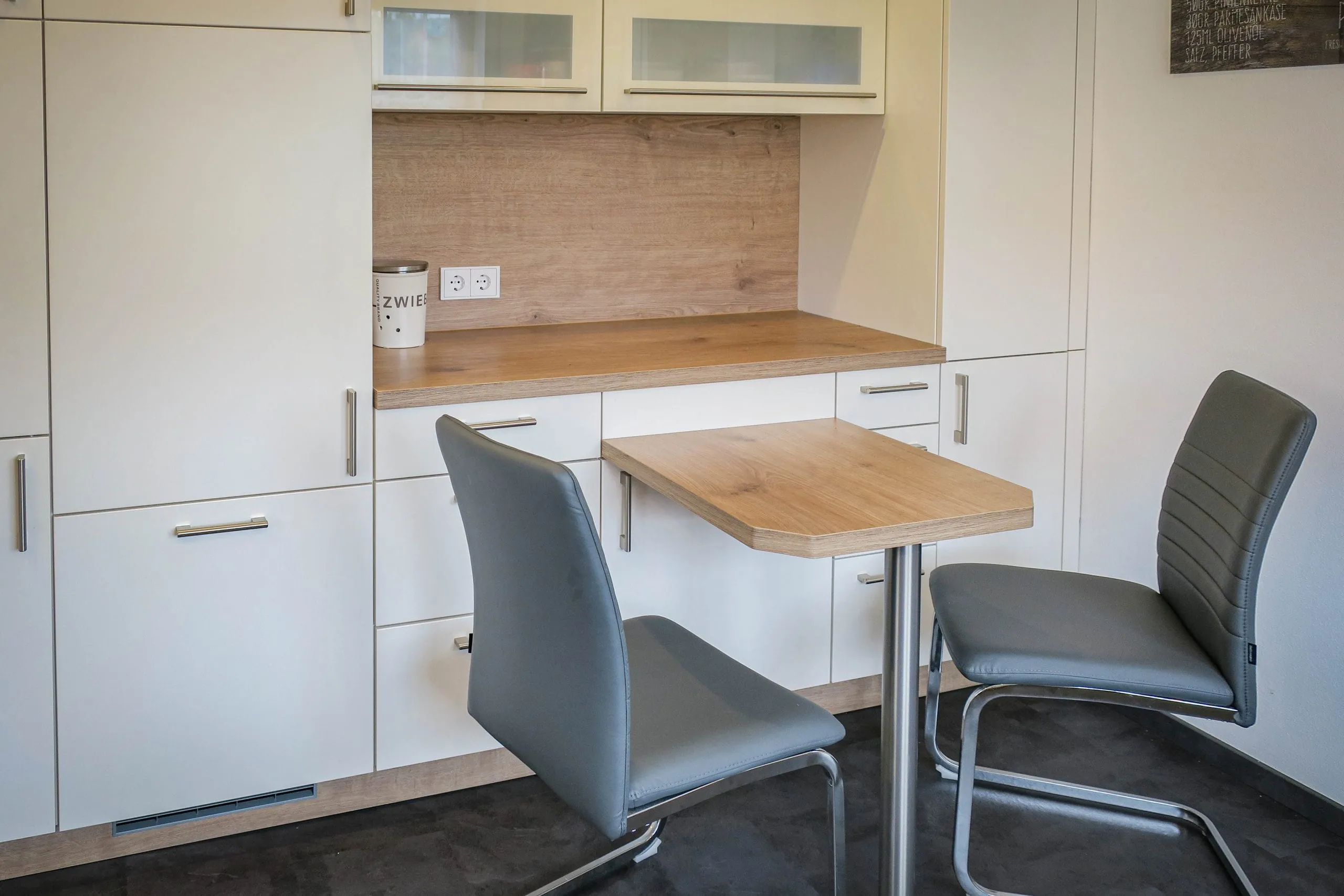 In Küchenzeile integrierter Esstisch in Holzoptik mit zwei grauen Lederstühlen, flankiert von weißen Hochschränken, mit Glas-Hängeschränken darüber