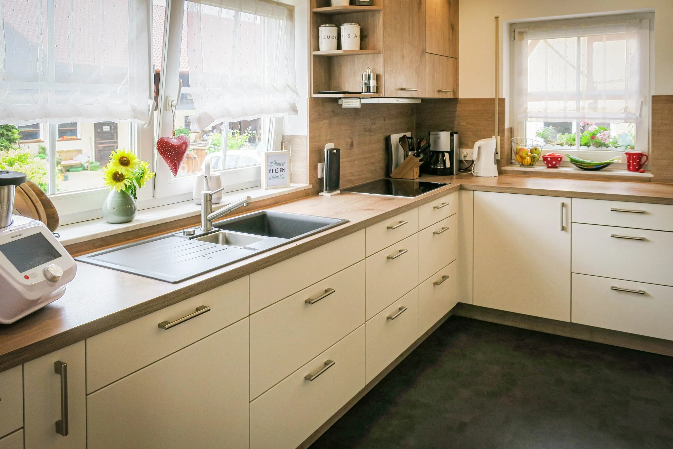 Eckküche um zwei Fenster geplant: Spüle am linken Fenster, Eckschrank mit Tellerbord, Holz-Arbeitsplatte und Beige-Fronten, im Hintergrund bepflanztes Fensterbrett