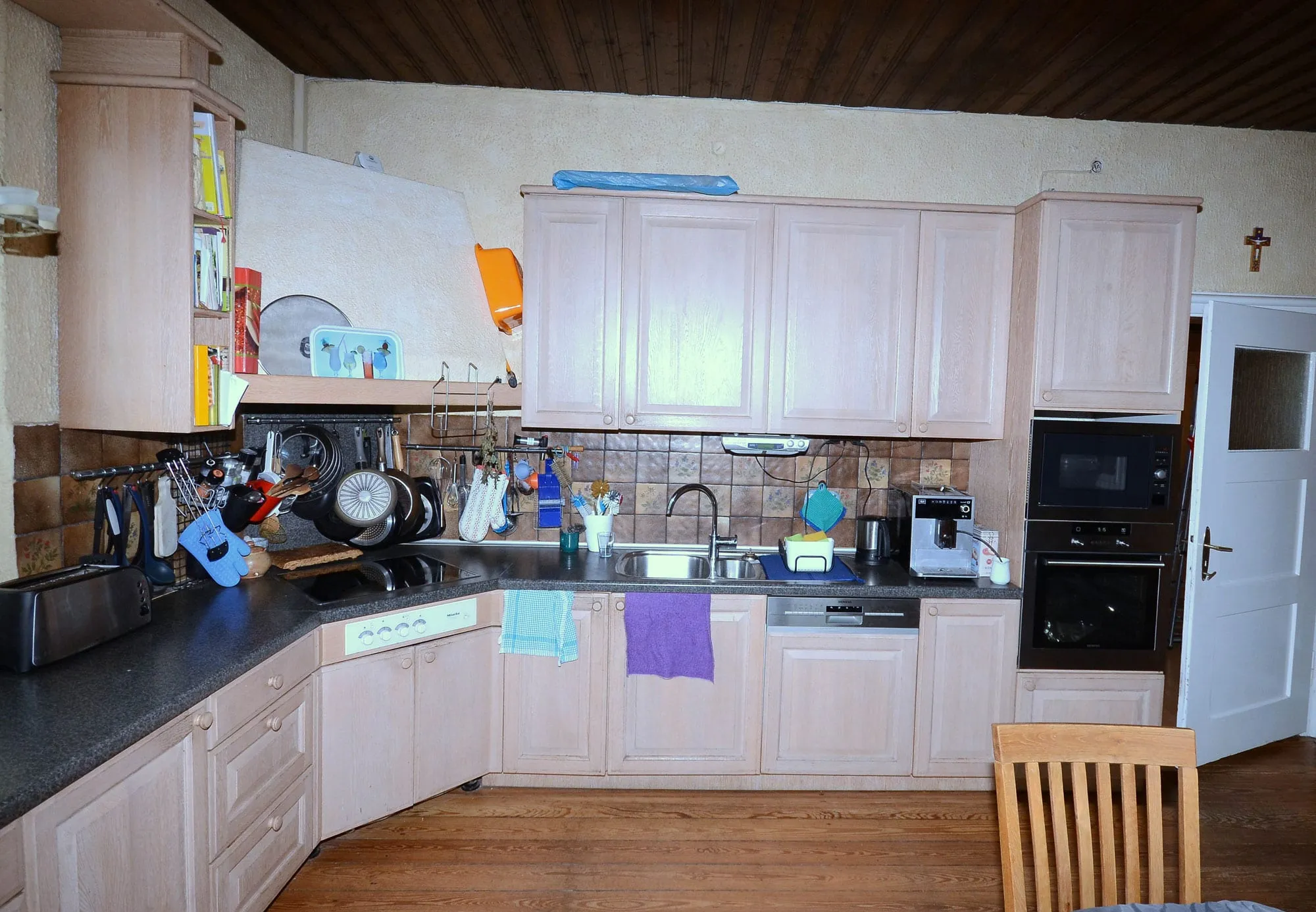 Vorher-Aufnahme einer L-förmigen Landhausküche mit hellen Holzfronten, gefliestem Spritzschutz und Holzbalkendecke – Ausgangssituation vor der Neuplanung durch Dietmar Kraus, Broßler Küche Aktiv
