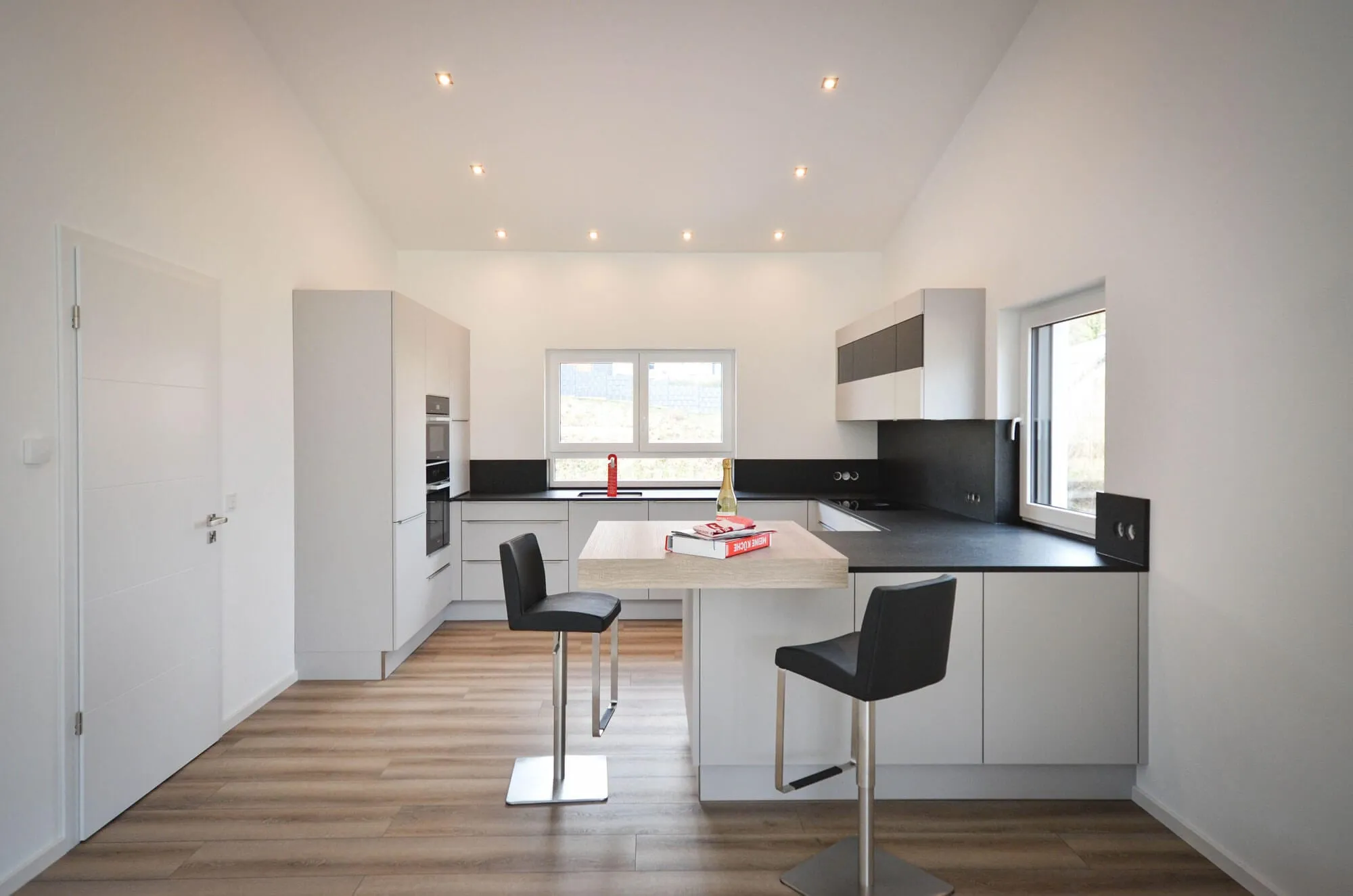 Moderne grifflose Küche in Hellgrau mit Kochinsel und Sitztheke aus Holz, zwei dunkle Barhocker, schwarze Arbeitsplatte und Hochschrankzeile mit integriertem Backofen, lichtdurchfluteter Raum mit hoher Decke und Eichendielen-Boden – Küchenplanung von Dietmar Kraus für Broßler Küche Aktiv