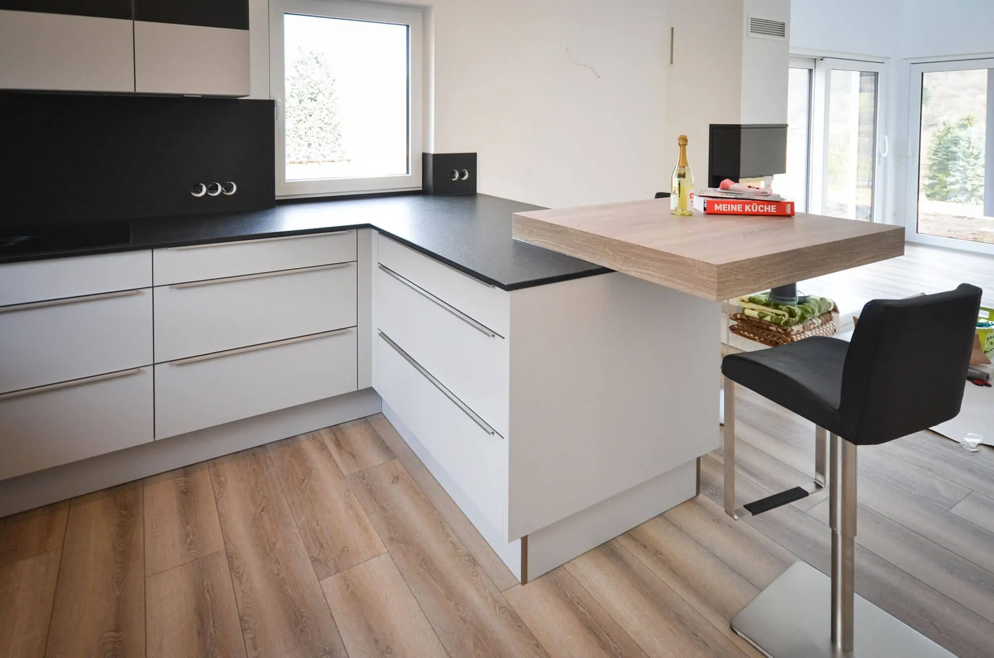 Moderne weiße Hochglanz-Küche in L-Form mit Kochinsel und integrierter Theke aus Holz, schwarze Arbeitsplatte, grifflose Fronten, lichtdurchfluteter Raum mit Eichendielen-Boden – Küchenplanung von Dietmar Kraus für Broßler Küche Aktiv