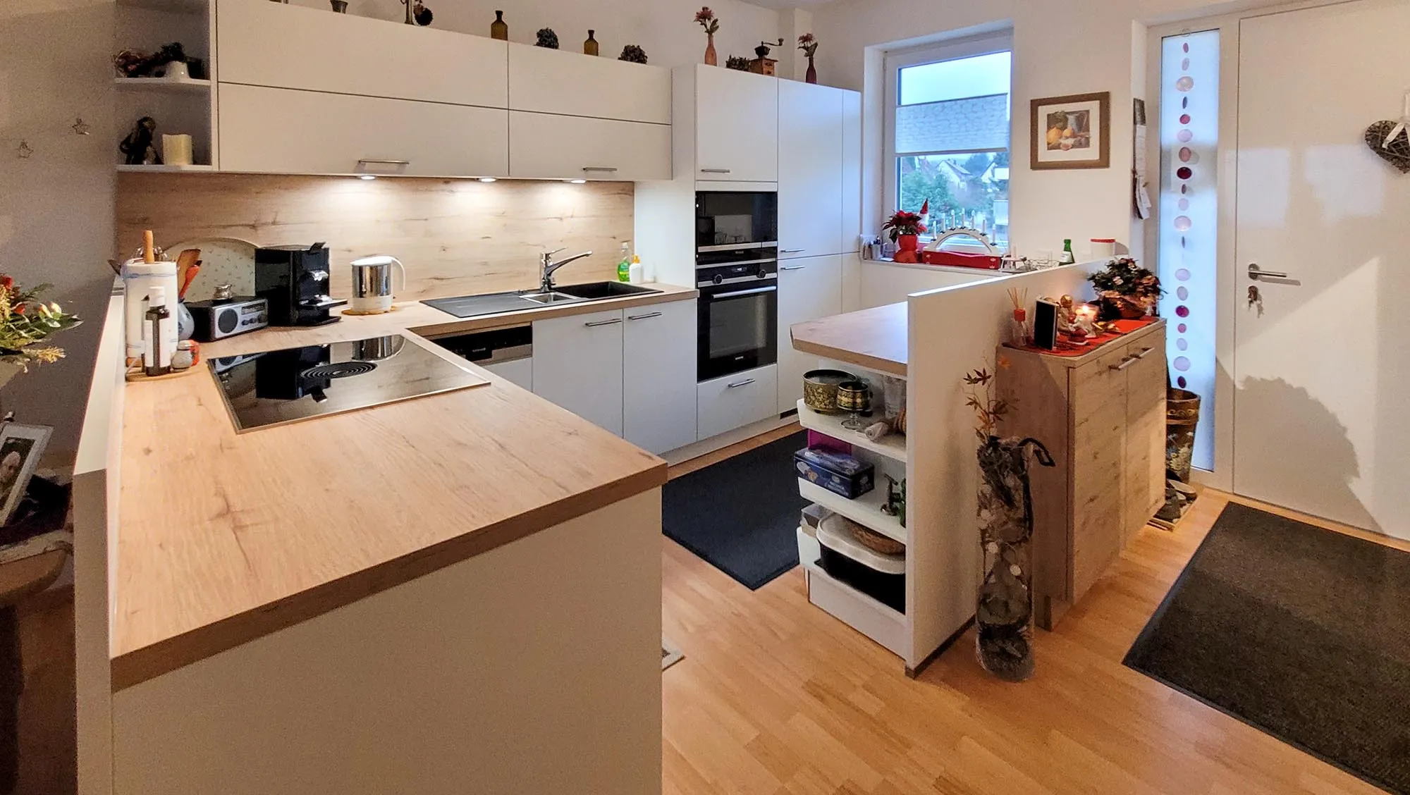 Vorher-Übersicht der bestehenden L-Küche mit weißen Fronten, Holz-Arbeitsplatte, Spüle am Fenster, schwarzem Backofen-Hochschrank, freistehendem Holz-Beistellschrank und Wandregalen