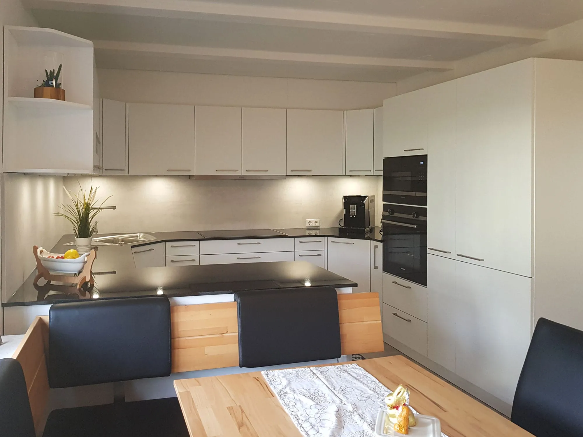 Magnolienfarbene L-Küche mit Halbinsel und integrierter Polsterbank, Backofen-Hochschrank rechts, im Vordergrund Esstisch mit Tischläufer