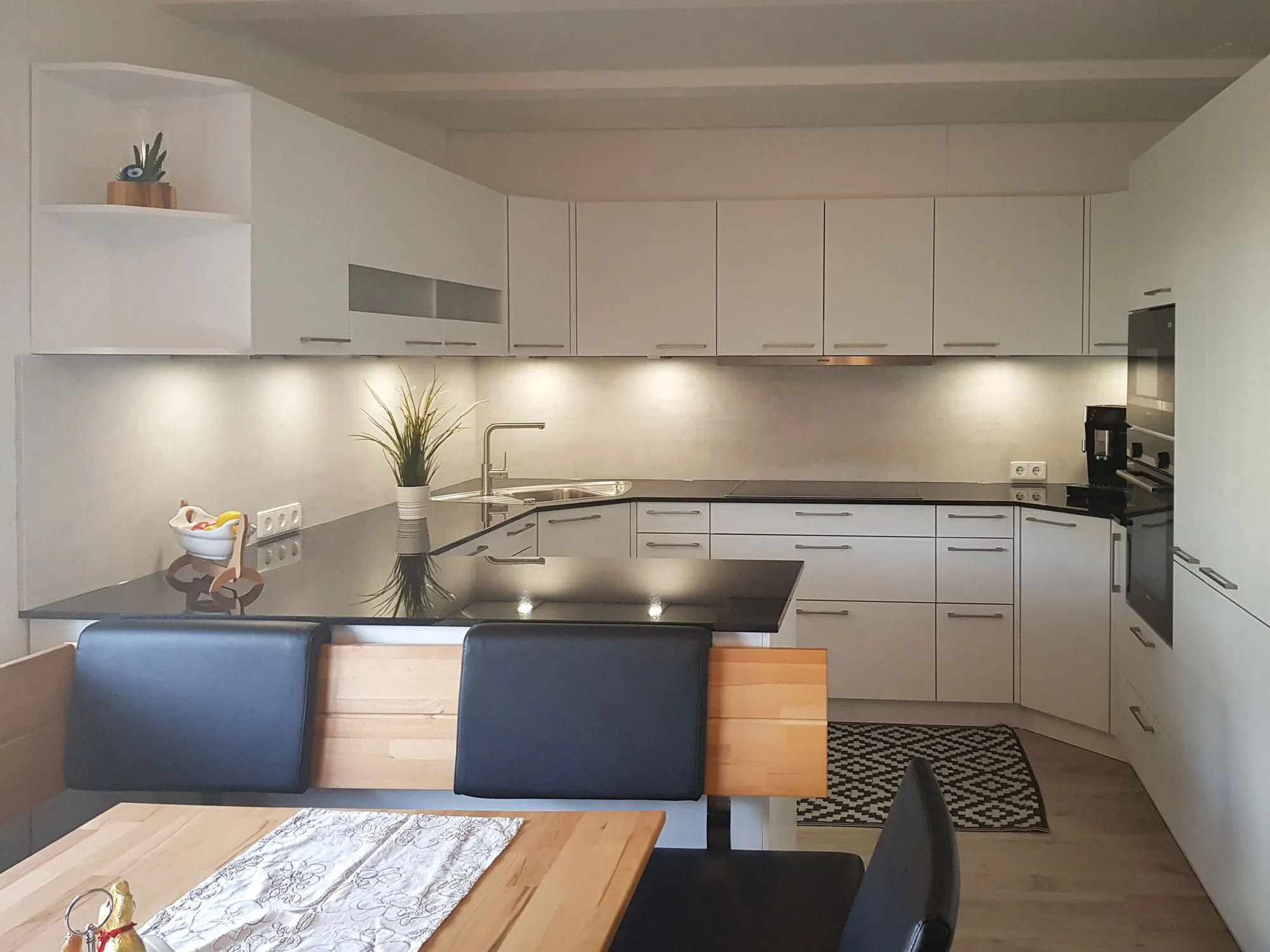 Blick über den Esstisch in die magnolienfarbene L-Küche mit Halbinsel: anthrazitfarbene Arbeitsplatte, Spüle, Backofen-Hochschrank und Eck-Tellerbord oben