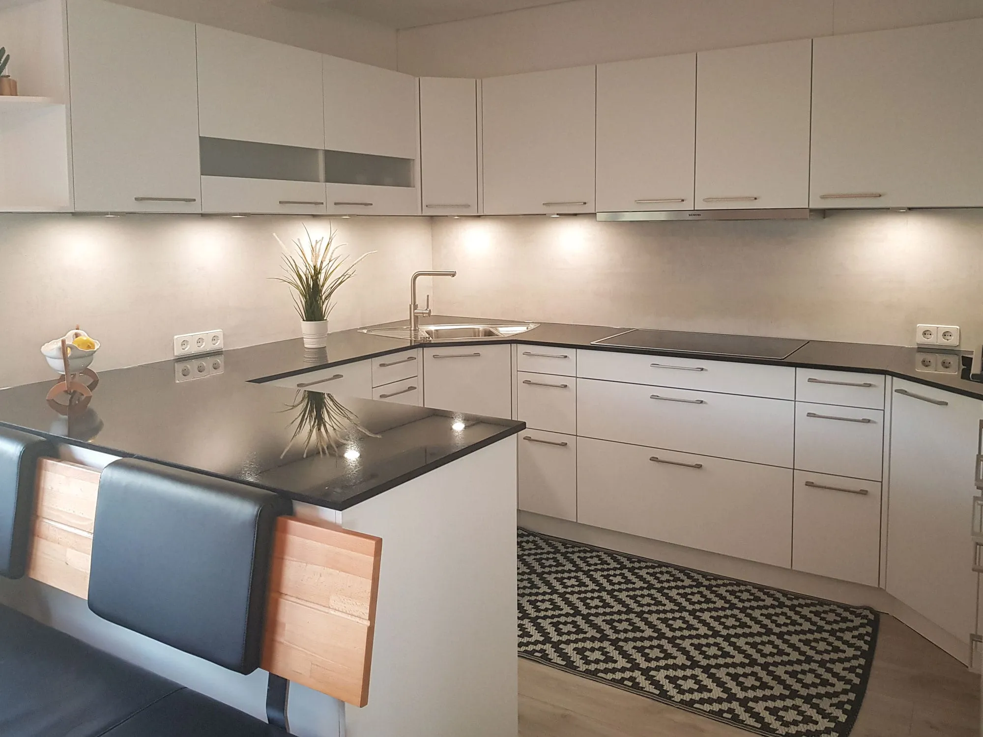 Magnolienfarbene L-Küche mit Halbinsel, anthrazitfarbener Arbeitsplatte, Spüle und Induktionskochfeld, im Vordergrund Halbinsel mit gepolsterter Sitzbank in Holz
