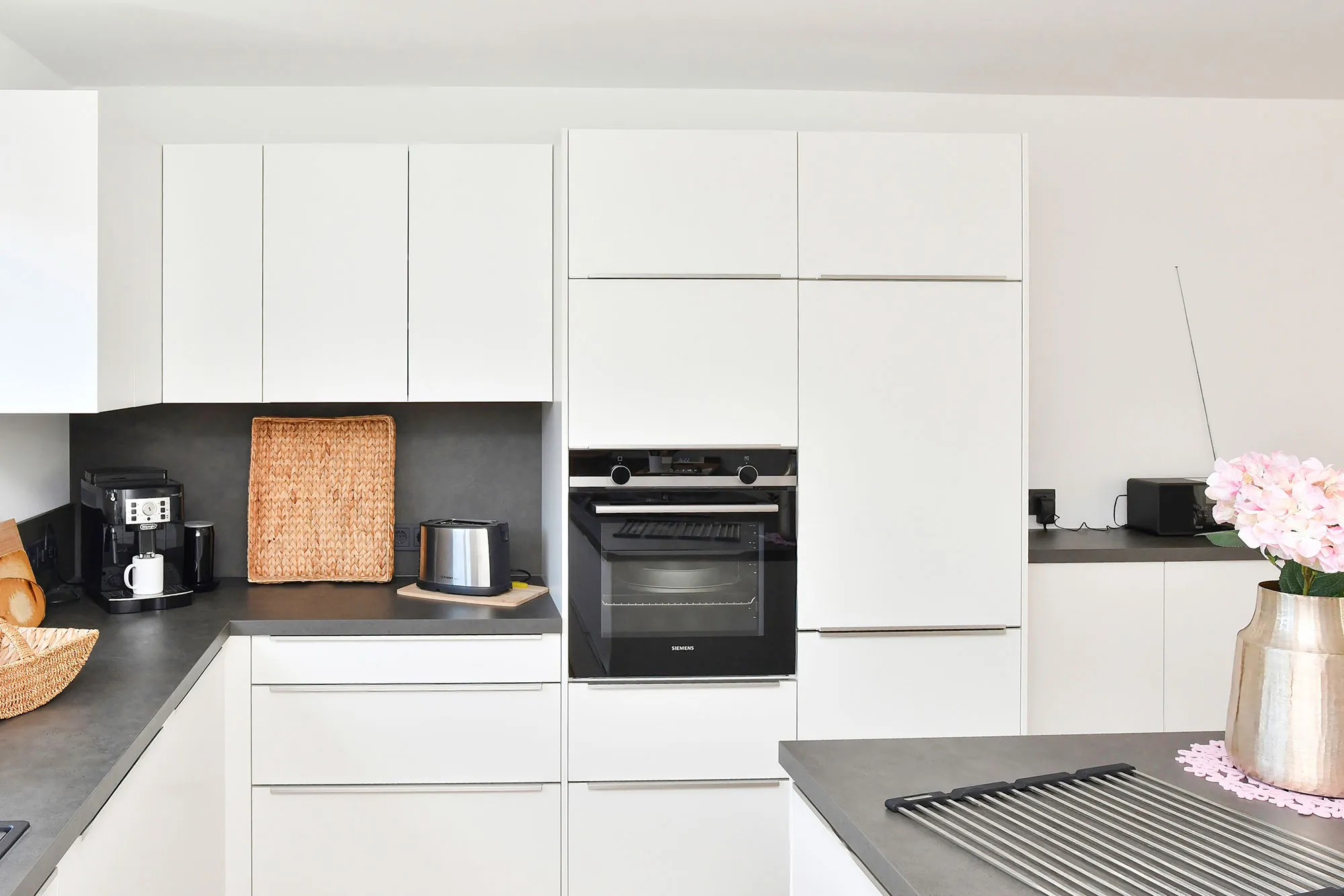 Weiße grifflose Küche mit raumhoher Hochschrankzeile, integriertem Backofen mittig und Kühlschrank rechts, dunkle Steinarbeitsplatte mit Toaster, Kaffeemaschine und Korb, im Vordergrund Kochinsel mit Muldenlüfter und Hortensienstrauß – Küchenplanung von Dietmar Kraus für Broßler Küche Aktiv