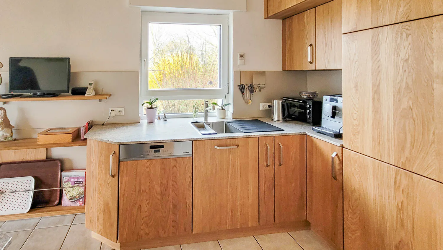 L-Hauptzeile in heller Buche/Erle mit Spüle vor Fenster und Hochschrank-Backofen rechts.