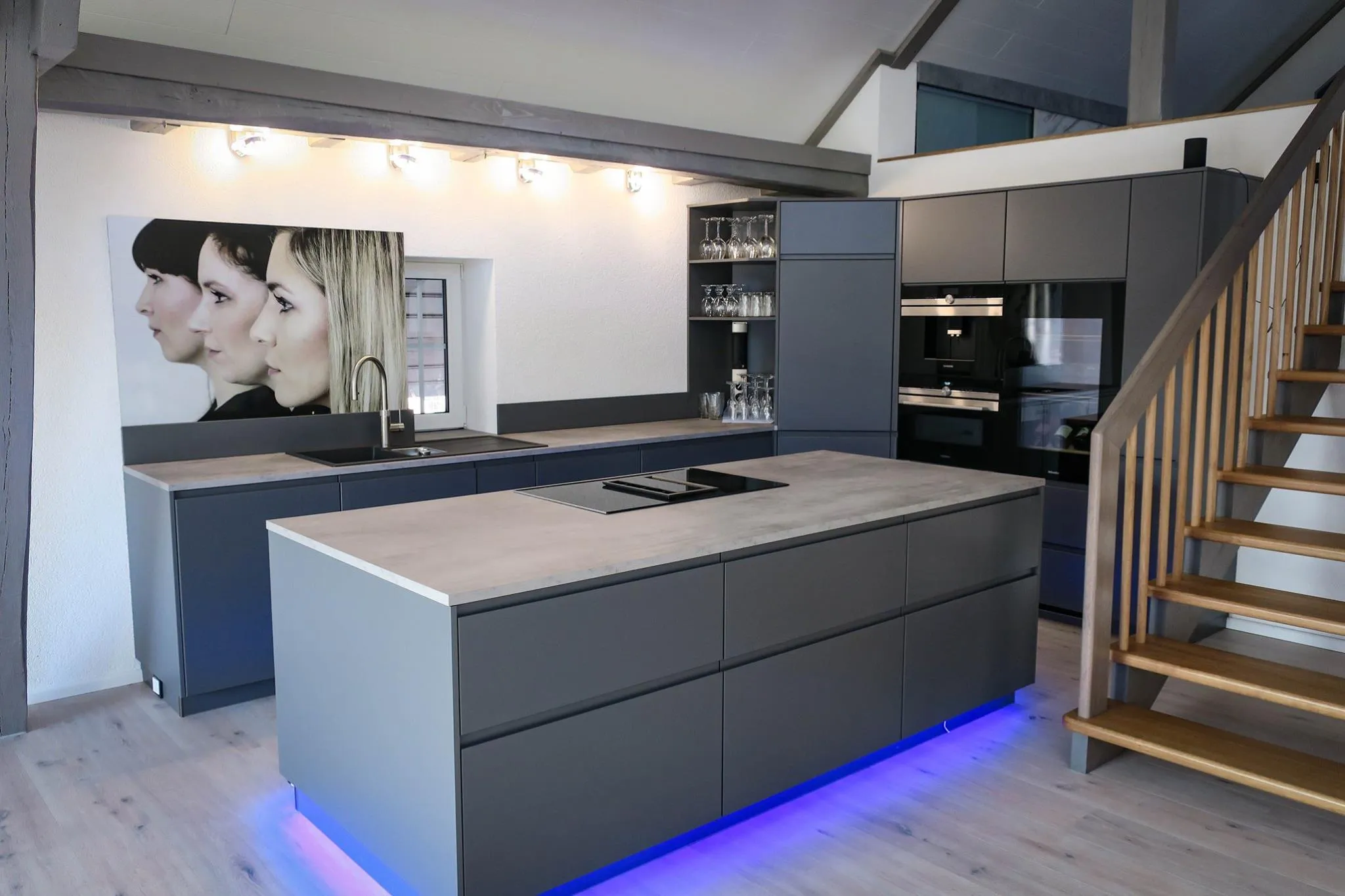 Gesamtansicht anthrazitfarbene Küche mit Kochinsel mit blauer LED-Sockelbeleuchtung, links Spülbereich mit großformatigem Schwarz-Weiß-Wandbild, rechts offene Holztreppe und Hochschrankzeile