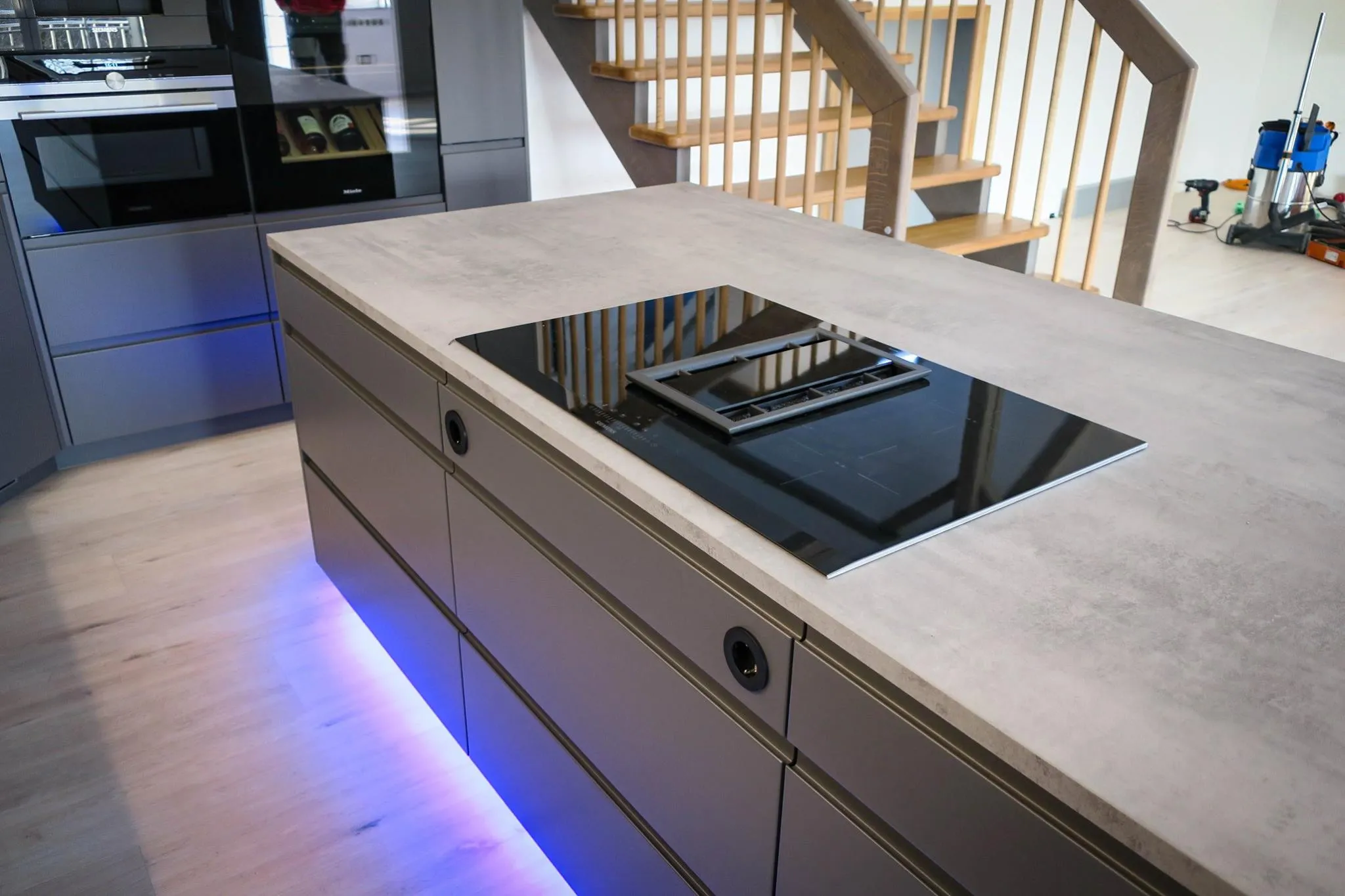 Detail Kochinsel mit Induktionskochfeld und integriertem Muldenlüfter, Stein-Arbeitsplatte in Beton-Optik und blauer LED-Sockelbeleuchtung, im Hintergrund Holztreppe und Hochschrank