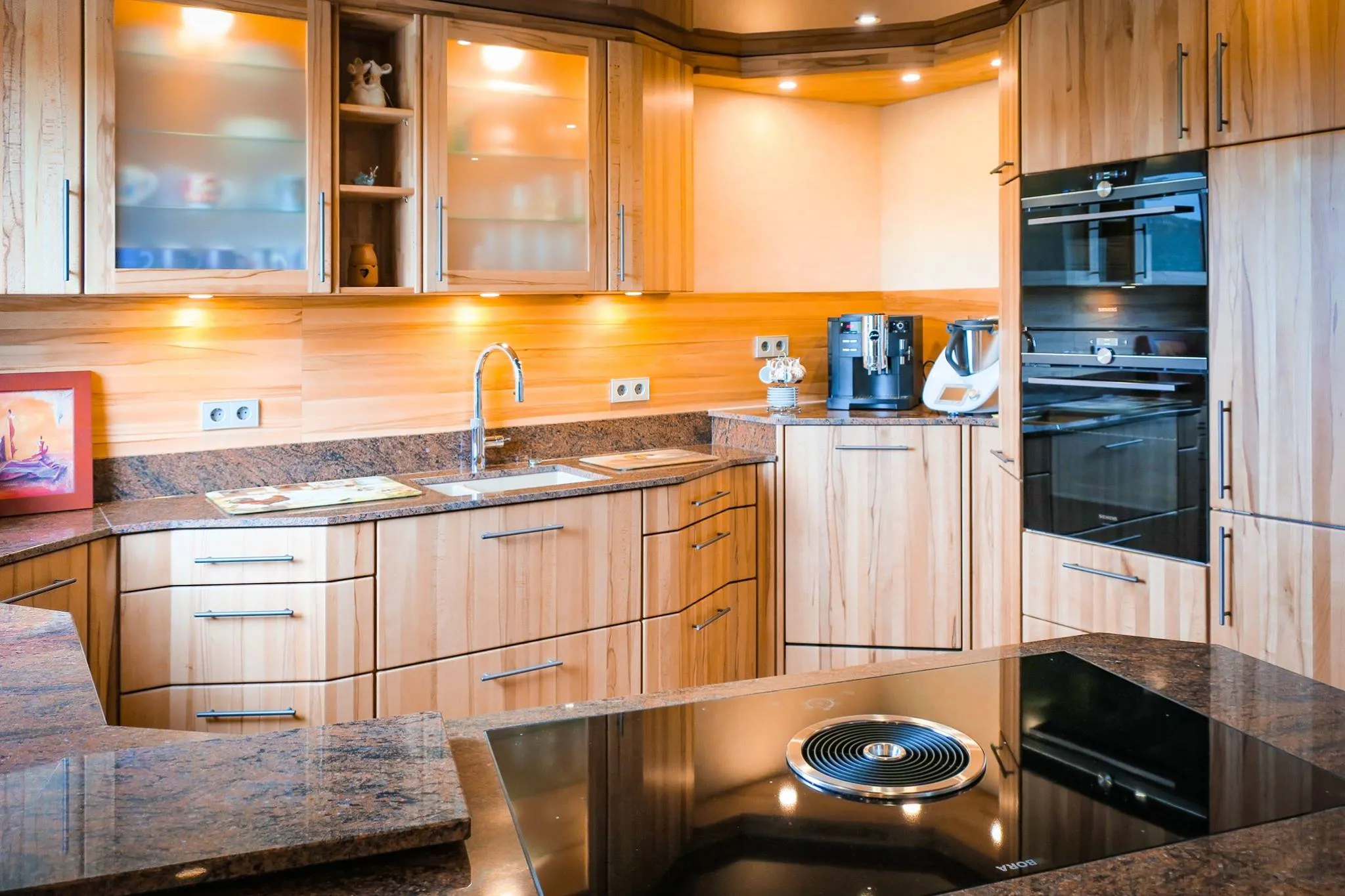 Eichen-Küche mit Backofen-Dampfgarer-Hochschrankzeile, Eck-Spülbereich mit Granit, Kaffeevollautomat, im Vordergrund Kochinsel mit Glaskeramikfeld und Granit-Arbeitsplatte