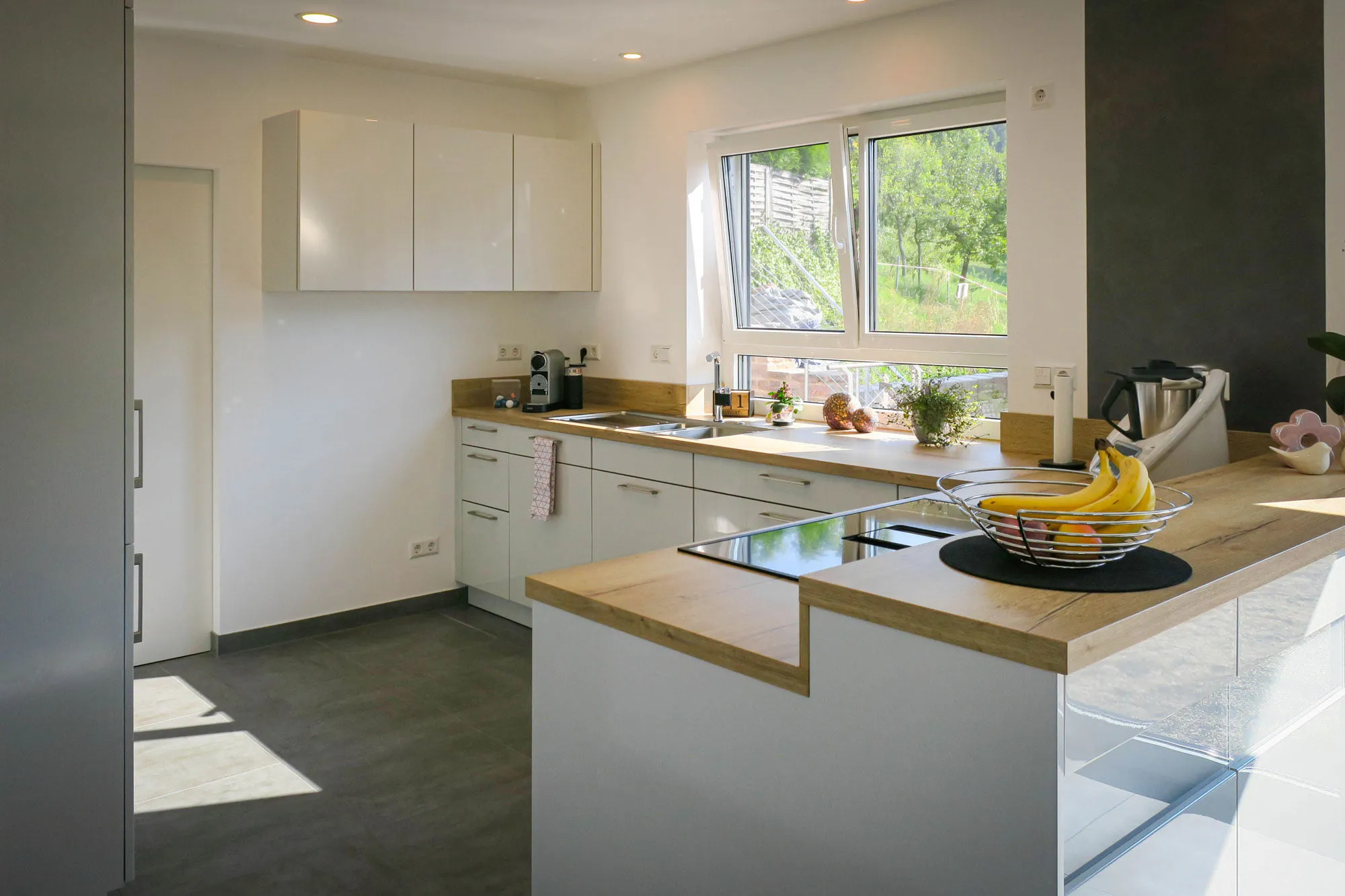 Helle weiße Küche mit Halbinsel und Holz-Bartheke vorne, Glaskeramik-Kochfeld, Spüle am Fenster mit Garten-Blick, Kühlschrank-Hochschrank links und Steinoptik-Fliesenboden