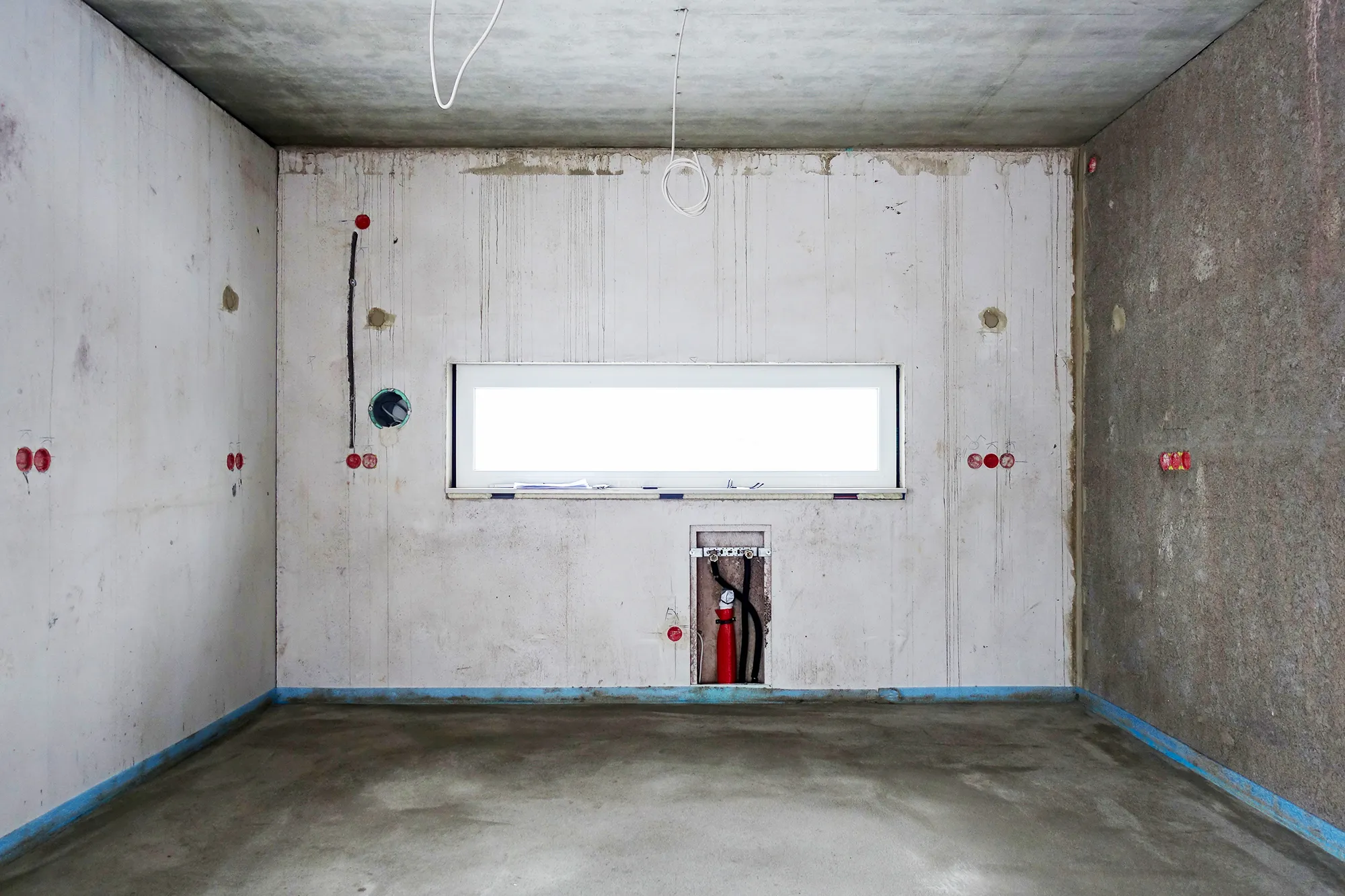 Vorher-Aufnahme der Rückwand im Rohbau vor dem Kücheneinbau, langes horizontales Fenster in der Wandmitte, sichtbare Vorinstallationen für Wasser- und Stromanschlüsse, Betonboden mit blauem Klebeband-Rand – Ausgangssituation für eine Küchenplanung von Dietmar Kraus bei Broßler Küche Aktiv