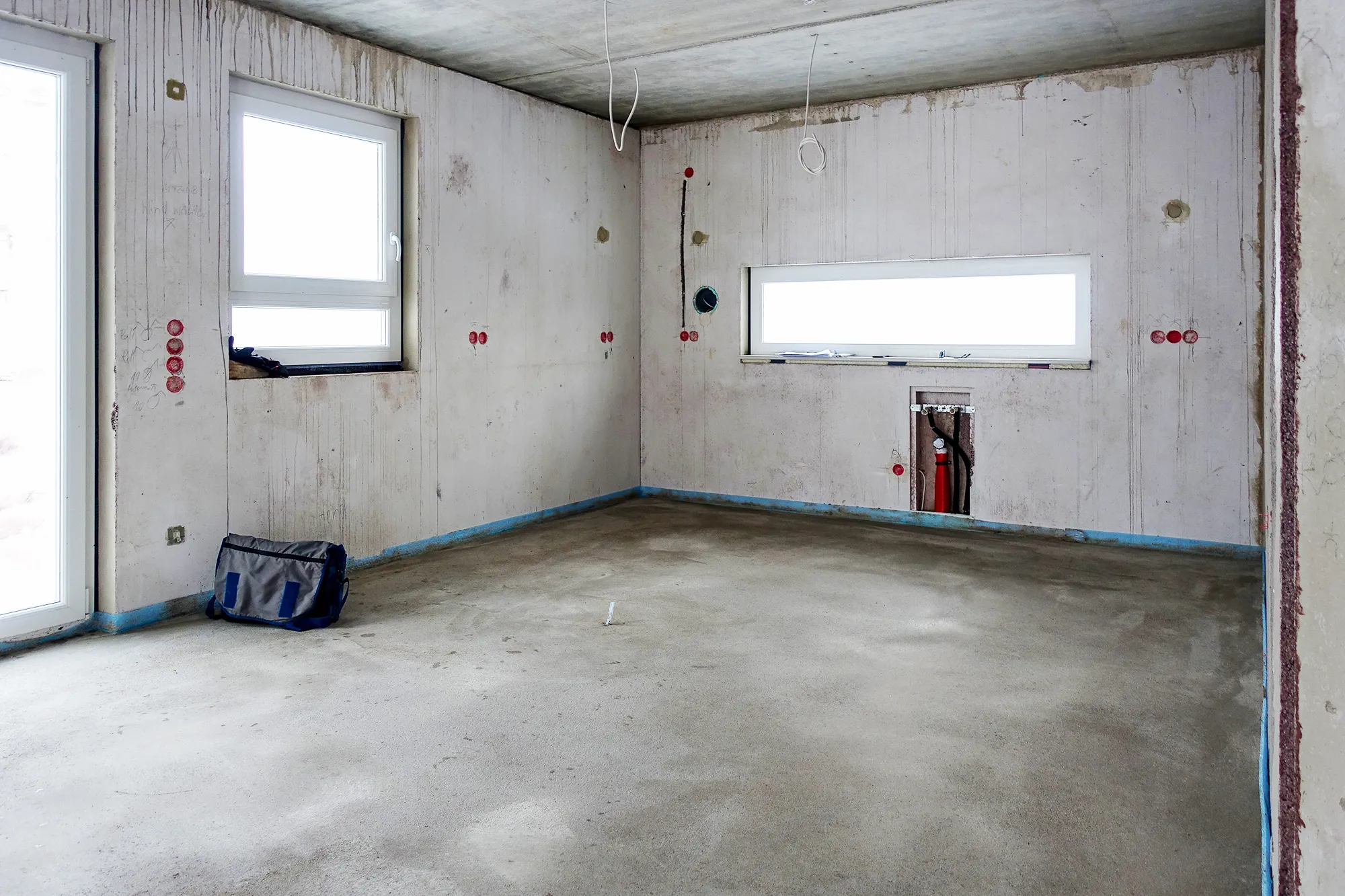 Leerer Rohbau-Raum vor dem Kücheneinbau, sichtbare Vorinstallationen für Wasser, Strom und Anschlüsse an den Wänden, zwei Fenster sorgen für Tageslicht, Betonboden mit Klebeband-Markierungen – Ausgangssituation für eine Küchenplanung von Dietmar Kraus bei Broßler Küche Aktiv