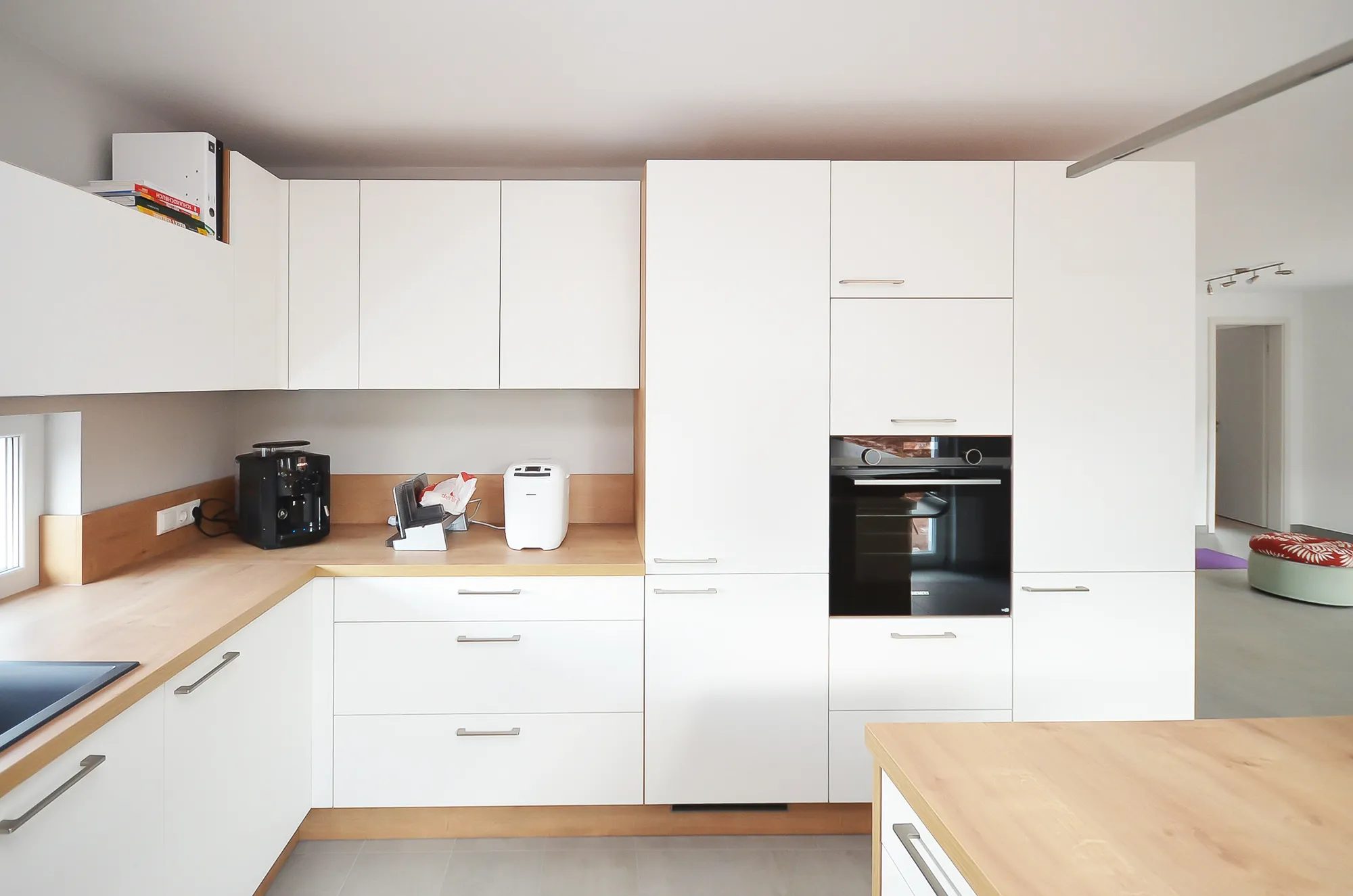 Weiße Küche mit raumhoher Hochschrankzeile und integriertem Backofen, Arbeitsfläche in Eichen-Holzoptik mit Kaffeevollautomat und Toaster, offener Übergang zum angrenzenden Wohnbereich – Küchenplanung von Dietmar Kraus für Broßler Küche Aktiv