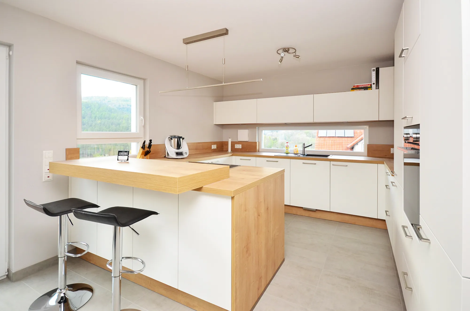 Helle U-Küche in Weiß mit vorgelagerter Sitztheke in Eichen-Holzoptik, zwei dunkle Barhocker mit Chromfuß, Fensterband über der Arbeitszeile mit Blick ins Grüne, dezente Beleuchtung über der Theke – Küchenplanung von Dietmar Kraus für Broßler Küche Aktiv