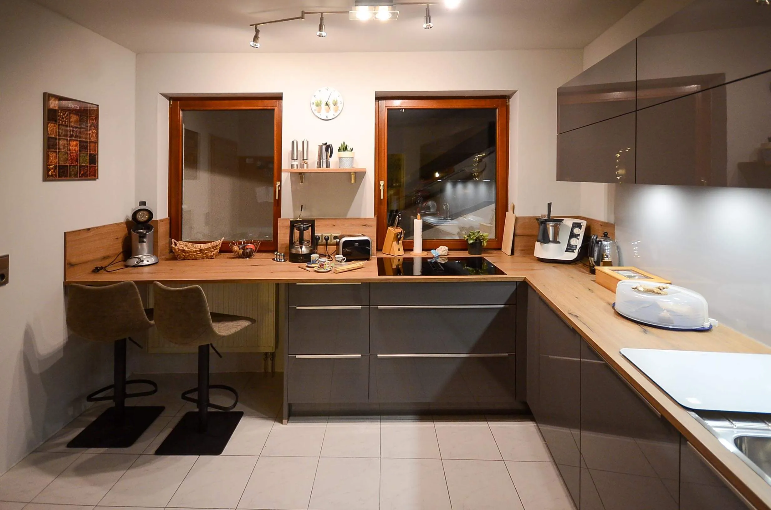 L-Küche mit dunkelbraunen Hochglanzfronten und Arbeitsplatte in Eichen-Holzoptik, integrierte Sitztheke mit zwei Designstühlen, Fenster mit altem Holzrahmen und Blick nach draußen, indirekte LED-Beleuchtung unter den Oberschränken – Küchenplanung von Dietmar Kraus für Broßler Küche Aktiv