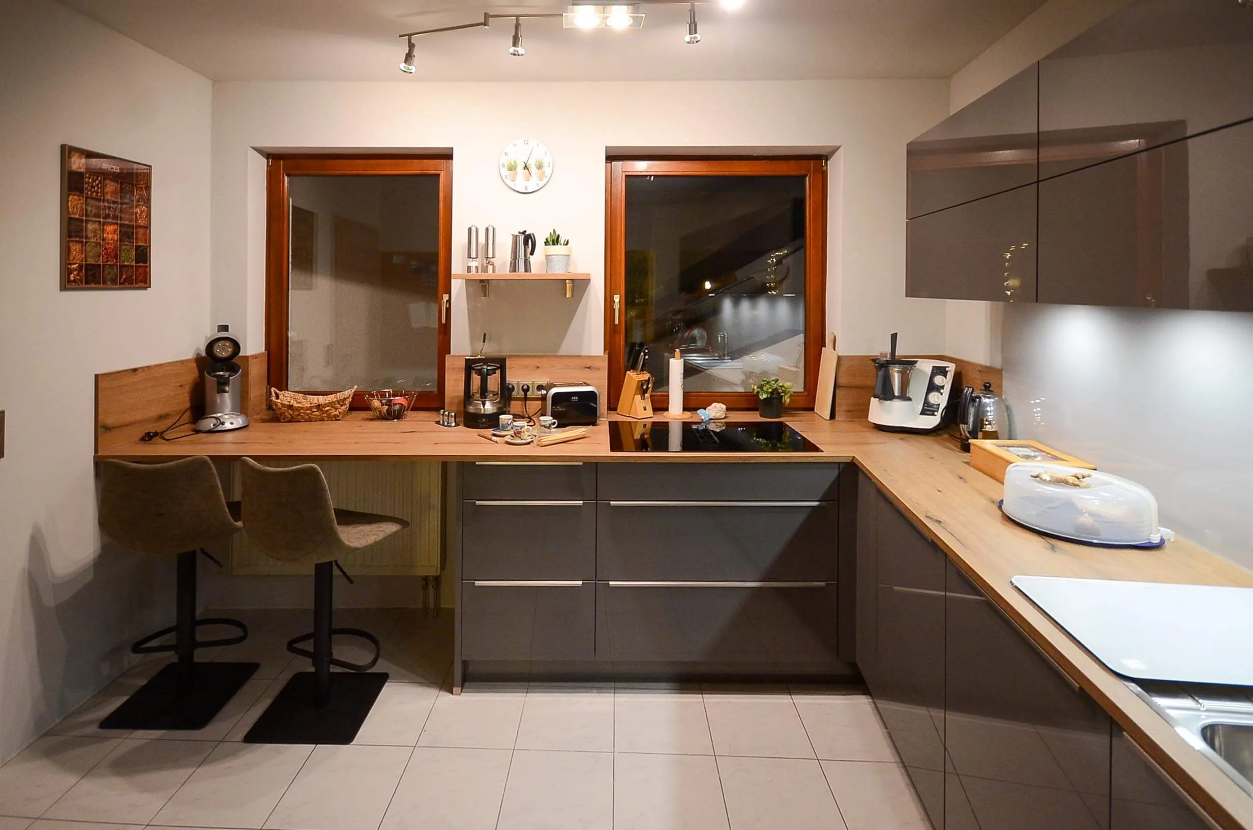 L-Küche mit dunkelbraunen Hochglanzfronten und Arbeitsplatte in Eichen-Holzoptik, integrierte Sitztheke mit zwei Designstühlen, Fenster mit altem Holzrahmen und Blick nach draußen, indirekte LED-Beleuchtung unter den Oberschränken – Küchenplanung von Dietmar Kraus für Broßler Küche Aktiv