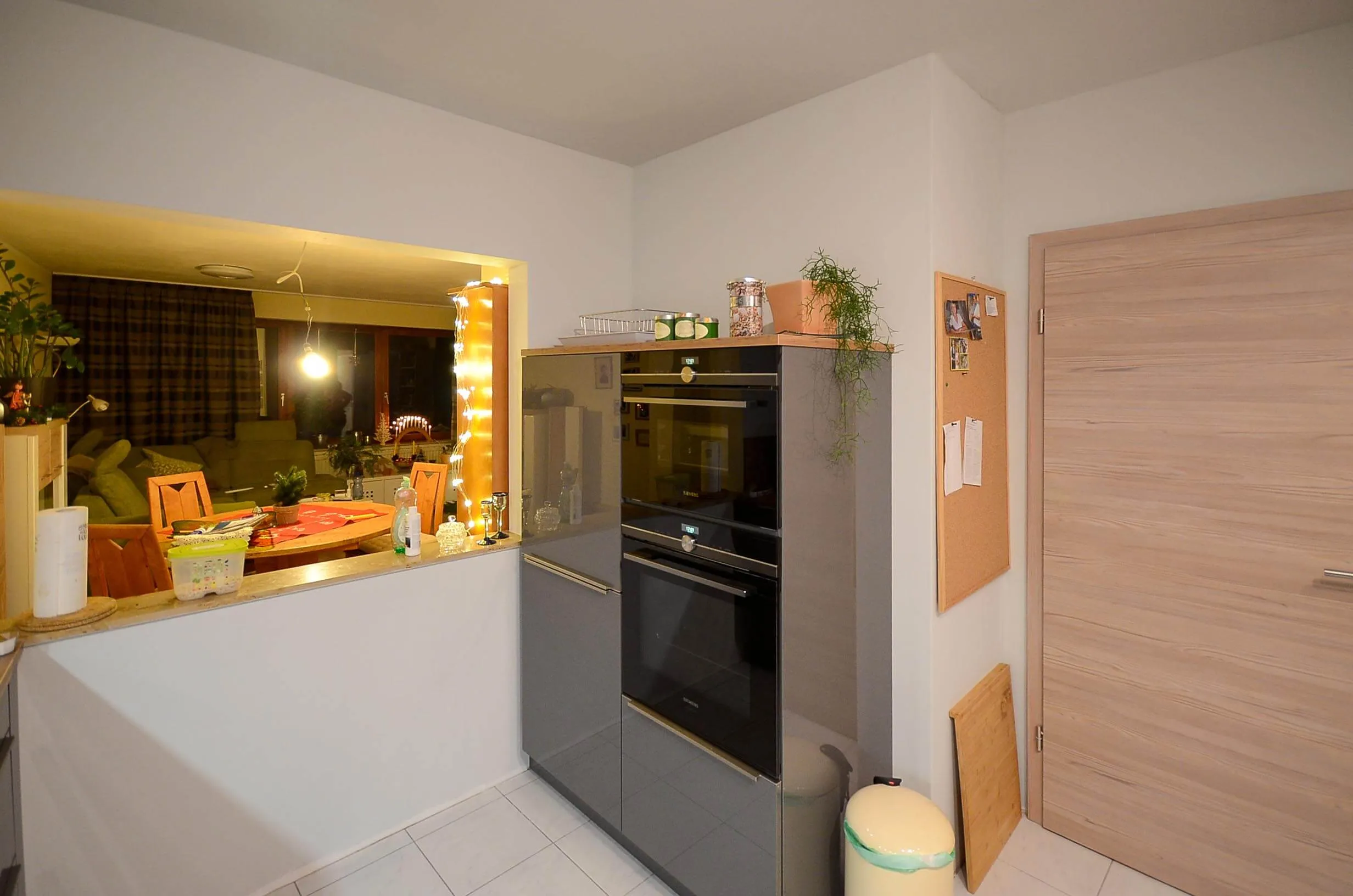 Seitliche Ansicht der dunkelbraunen Hochglanz-Küche mit raumhohem Backofenturm samt integriertem Backofen und Mikrowelle, links Durchreiche mit Blick ins angrenzende Esszimmer mit warmer Lichterkette, rechts Tür in Eichenoptik – Küchenplanung von Dietmar Kraus für Broßler Küche Aktiv