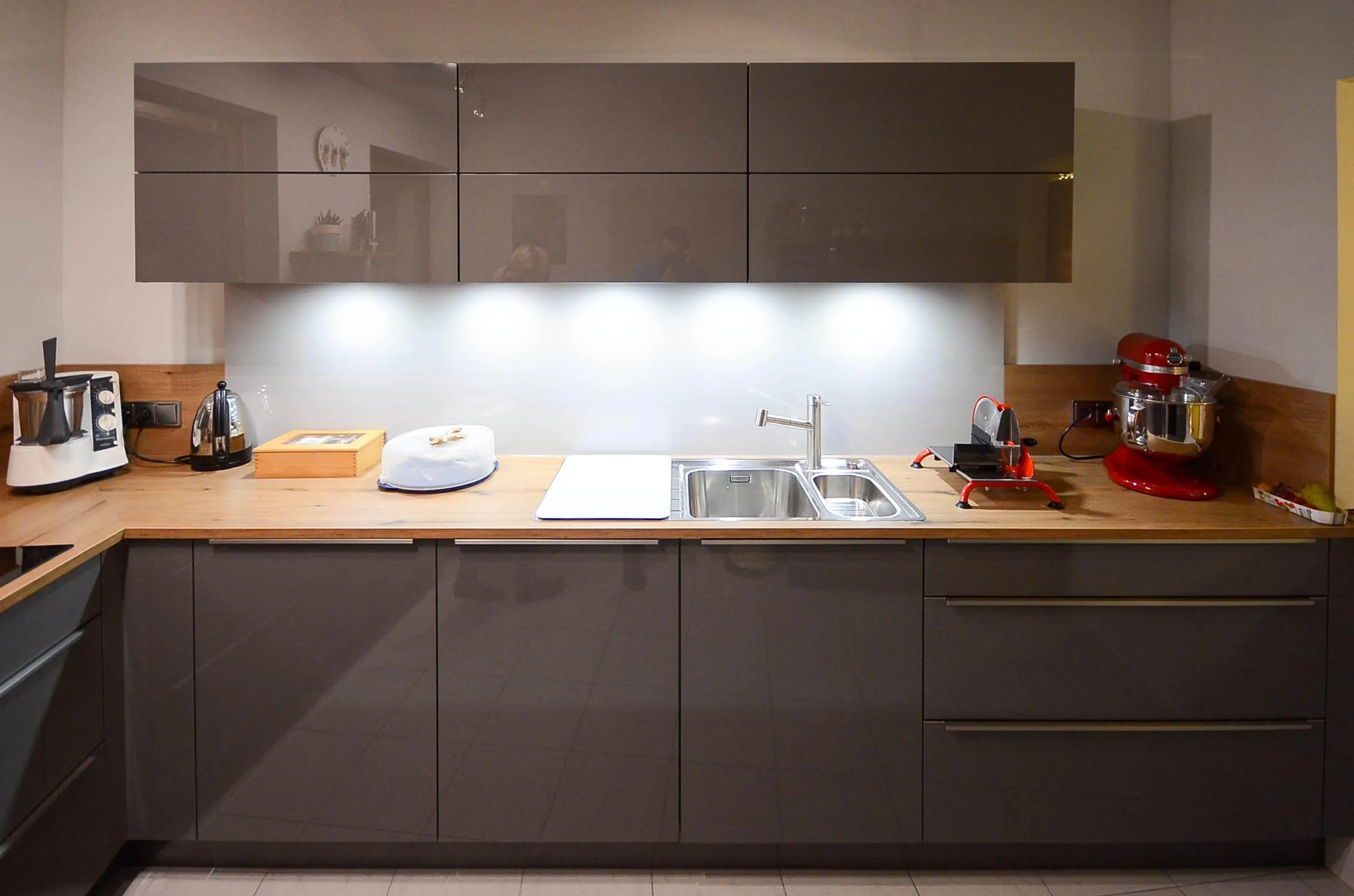 Frontaler Blick auf die Arbeitszeile der Küche mit dunkelbraunen Hochglanzfronten und Eichen-Arbeitsplatte, doppelte Edelstahl-Spüle vor heller Rückwand mit indirekter Beleuchtung, rote KitchenAid-Küchenmaschine als Farbakzent rechts – Küchenplanung von Dietmar Kraus für Broßler Küche Aktiv