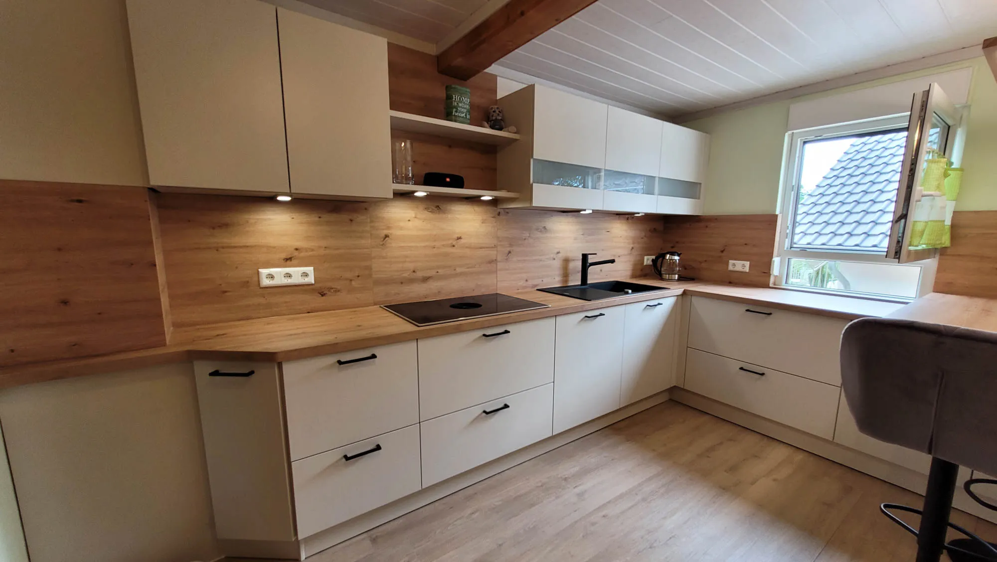 U-Küche mit Holzbalken-Decke, beigen grifflosen Fronten, Holz-Rückwand und Holz-Arbeitsplatte, schwarzem Wand-Dunstabzug über Induktionskochfeld und integrierter Bartheke am Fenster