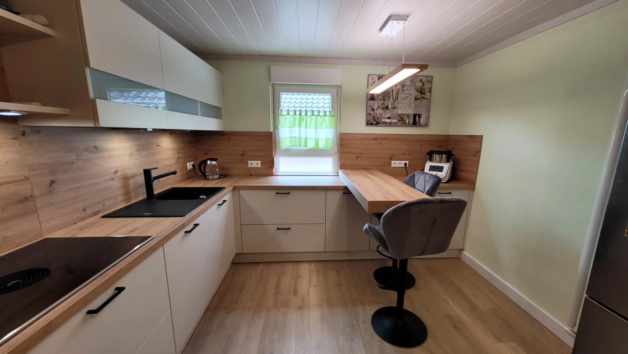 U-Küche mit beigen grifflosen Fronten, Holz-Arbeitsplatte und Holz-Rückwand, schwarzer Spüle und Armatur, Induktionskochfeld links und integriertem Holz-Tresen unter dem Fenster