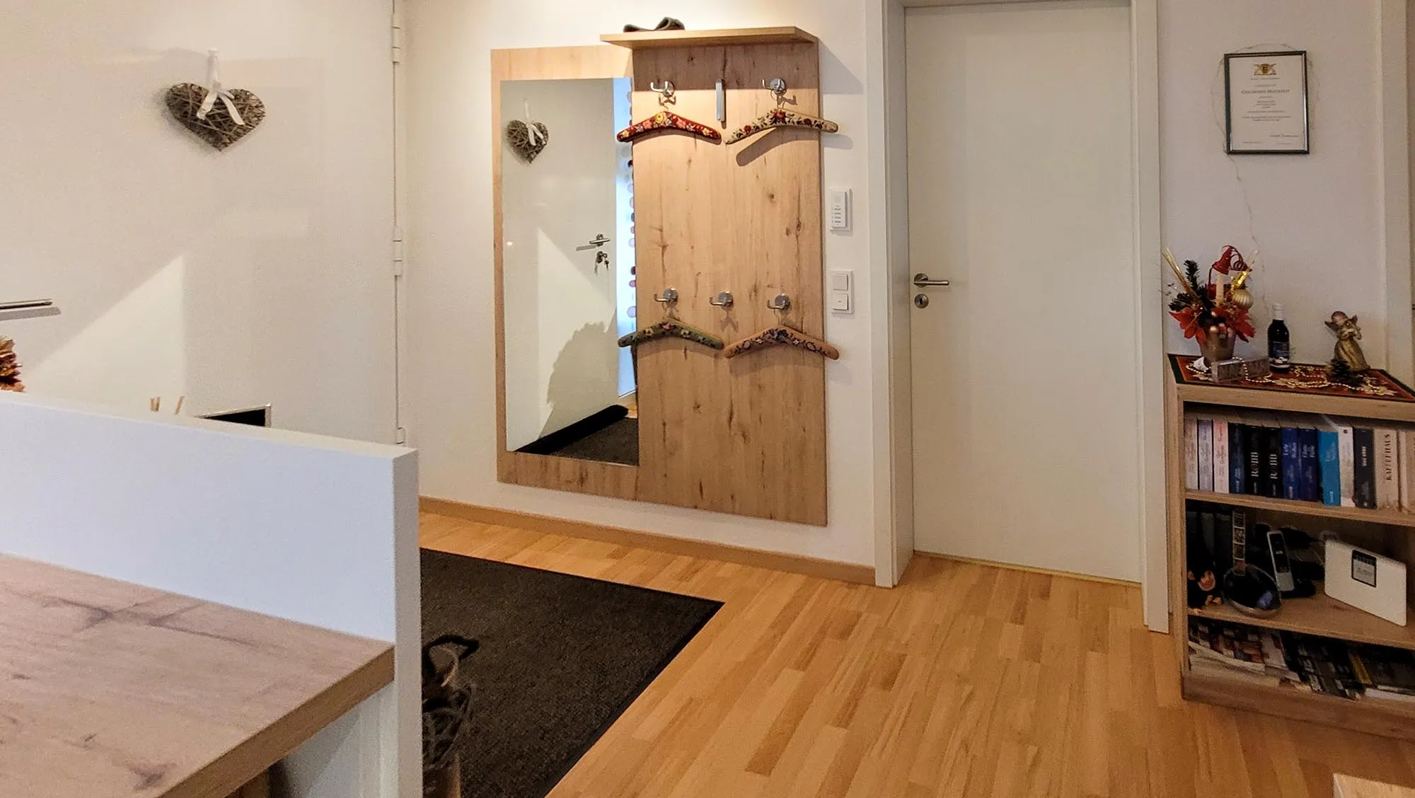Vorher-Aufnahme des Eingangs- und Garderobenbereichs neben der Küche: Holzgarderobe mit Spiegel, Bücherregal rechts, links angeschnitten die bestehende Holz-Arbeitsplatte der Bestandsküche