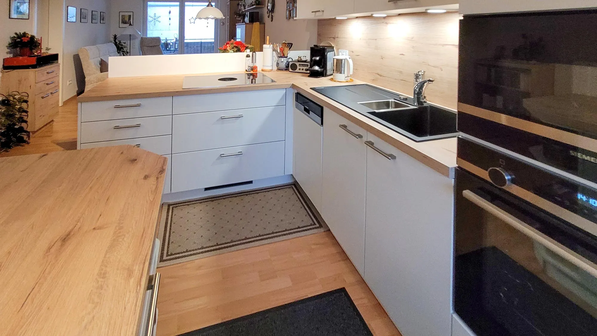 Vorher-Aufnahme der bestehenden L-Küche mit weißen Fronten, Holz-Arbeitsplatte, Spüle und schwarzem Backofen-Hochschrank, Weitwinkel-Aufnahme zur Raumdokumentation