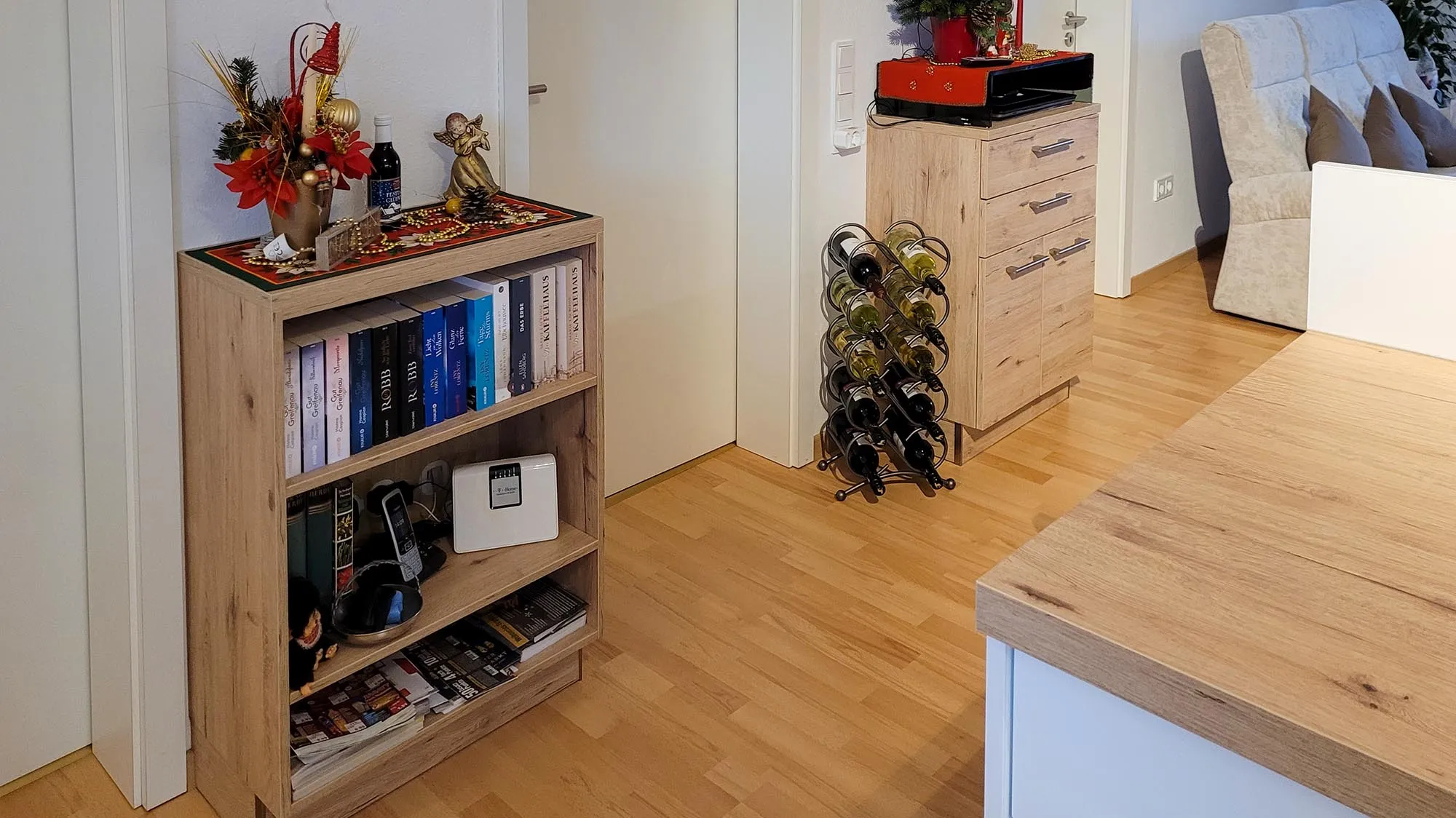 Vorher-Aufnahme des Raums vor dem Küchenumbau: Holzboden, Bücherregal und Weinregal an der Wand, rechts ein Sessel und im Vordergrund die bestehende Holz-Arbeitsplatte