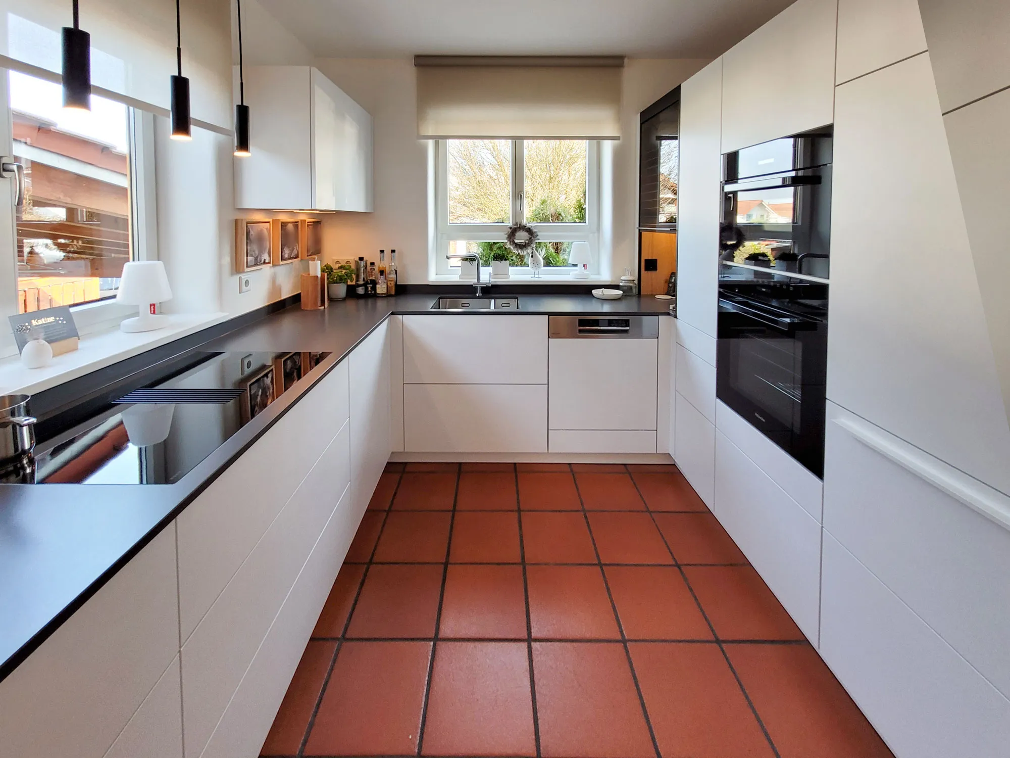 Frontalansicht der weißen U-Küche mit Spüle am Fenster mittig, Backofen-Hochschrank rechts, schwarzen Pendelleuchten, anthrazitfarbener Arbeitsplatte und terrakottafarbenem Fliesenboden