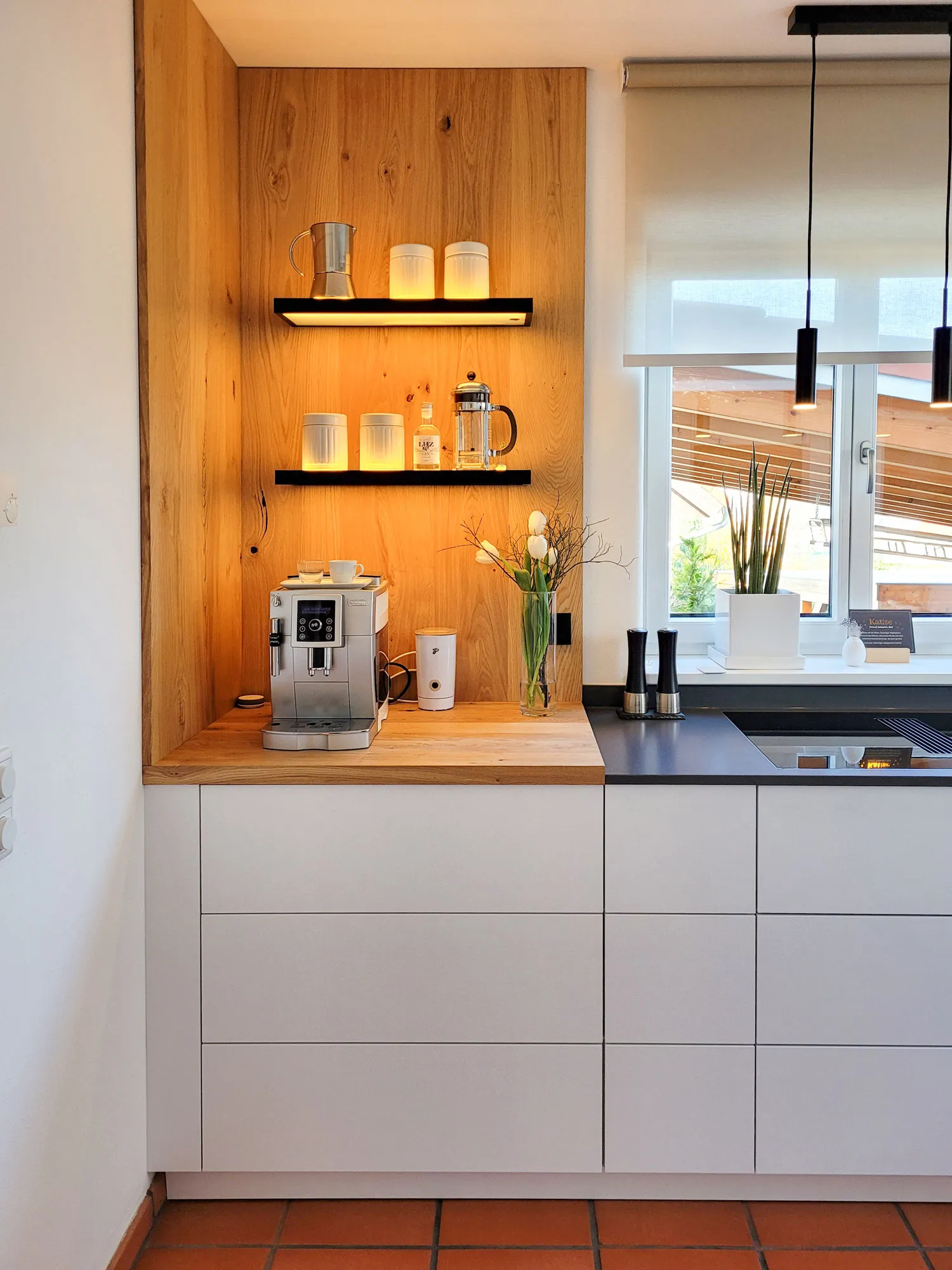 Holznische mit beleuchteten Holzregalen, Kaffeevollautomat auf Holz-Arbeitsplatte und Tulpen-Vase, rechts schwarze Pendelleuchten und Blick zur Küchenzeile am Fenster