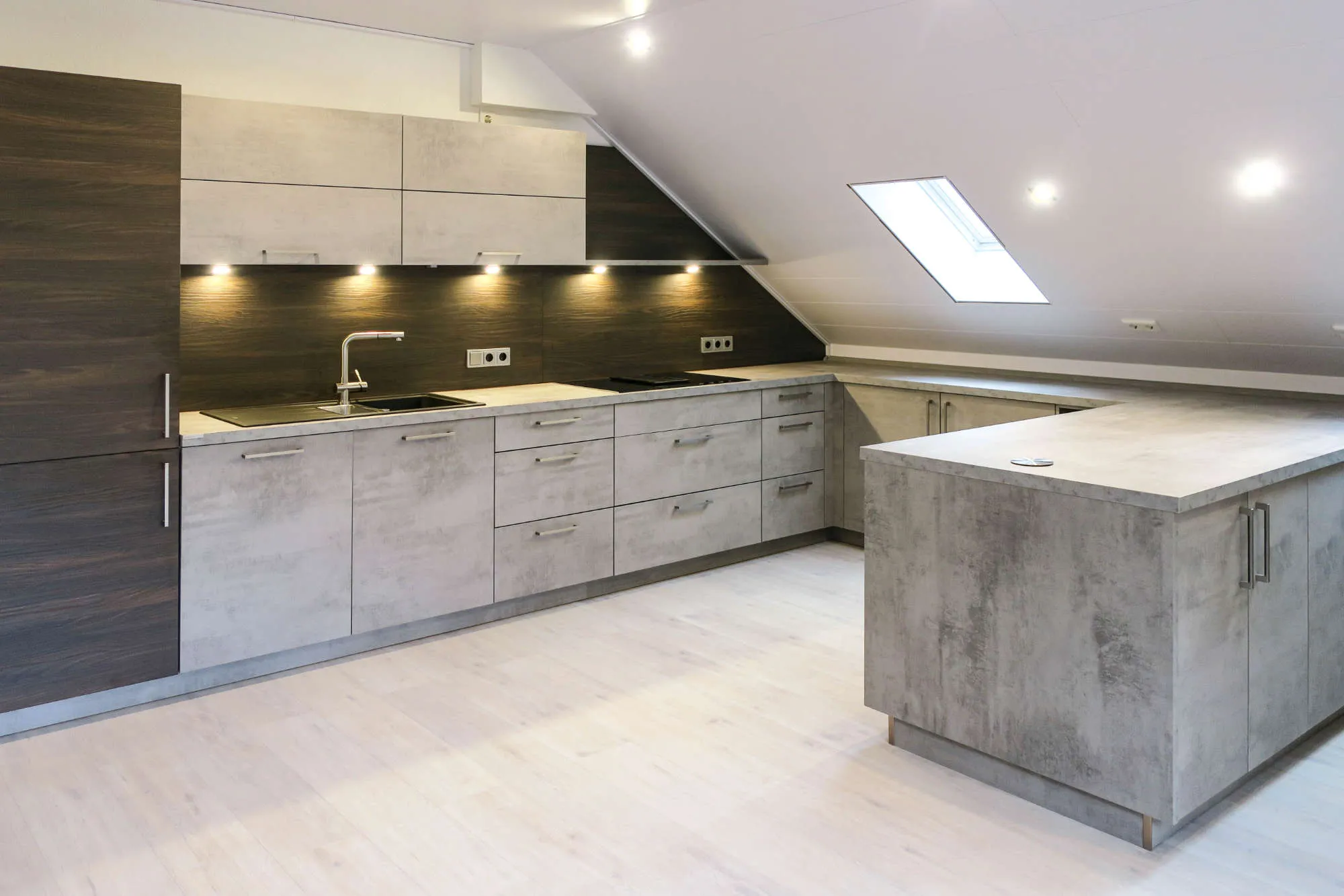 Gesamtansicht der fertigen Dachgeschossküche in L-Form mit Halbinsel: dunkle Holznische, Spüle und Induktionskochfeld an der Wand, grifflose Unterschränke und Halbinsel in Betonoptik unter der Dachschräge mit Dachfenster