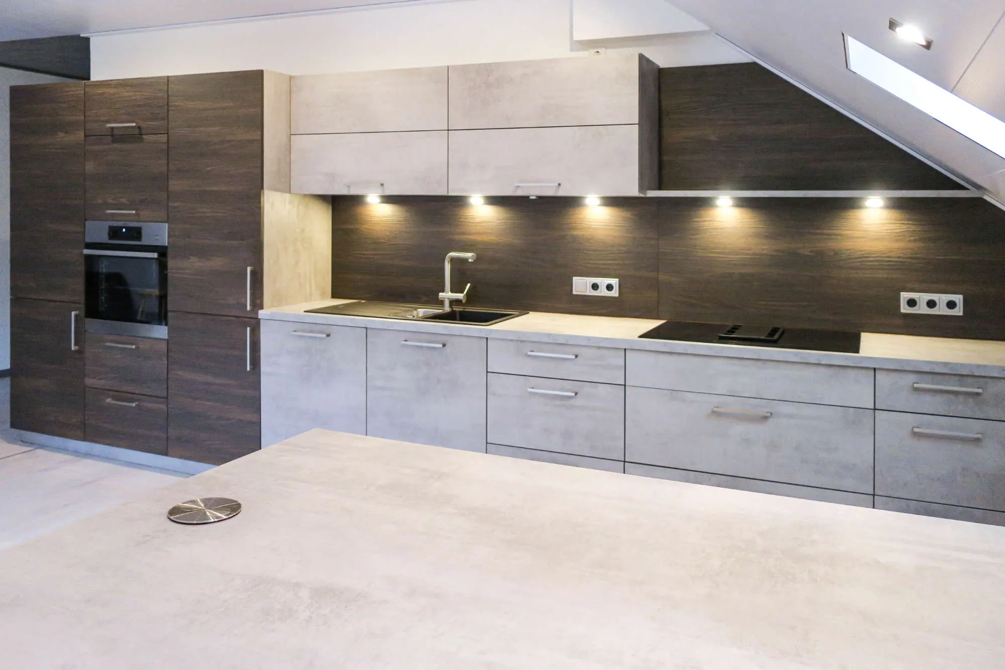 Dachgeschossküche mit dunkler Holznische und Betonoptik-Fronten, Backofen-Hochschrank, Spüle, Induktionskochfeld, Dachfenster über der Arbeitszeile und Kochinsel mit eingelassener Steckdose
