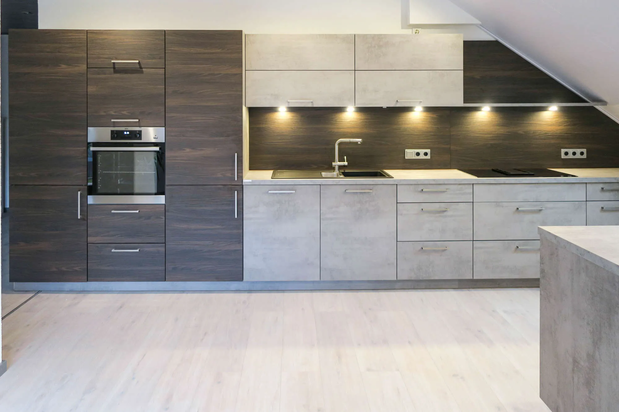 Moderne Dachgeschossküche unter Dachschräge mit dunklen Holz-Hochschränken, grifflosen Unterschränken in Betonoptik, integriertem Backofen-Hochschrank, Spüle und Induktionskochfeld sowie Kochinsel in Betonoptik