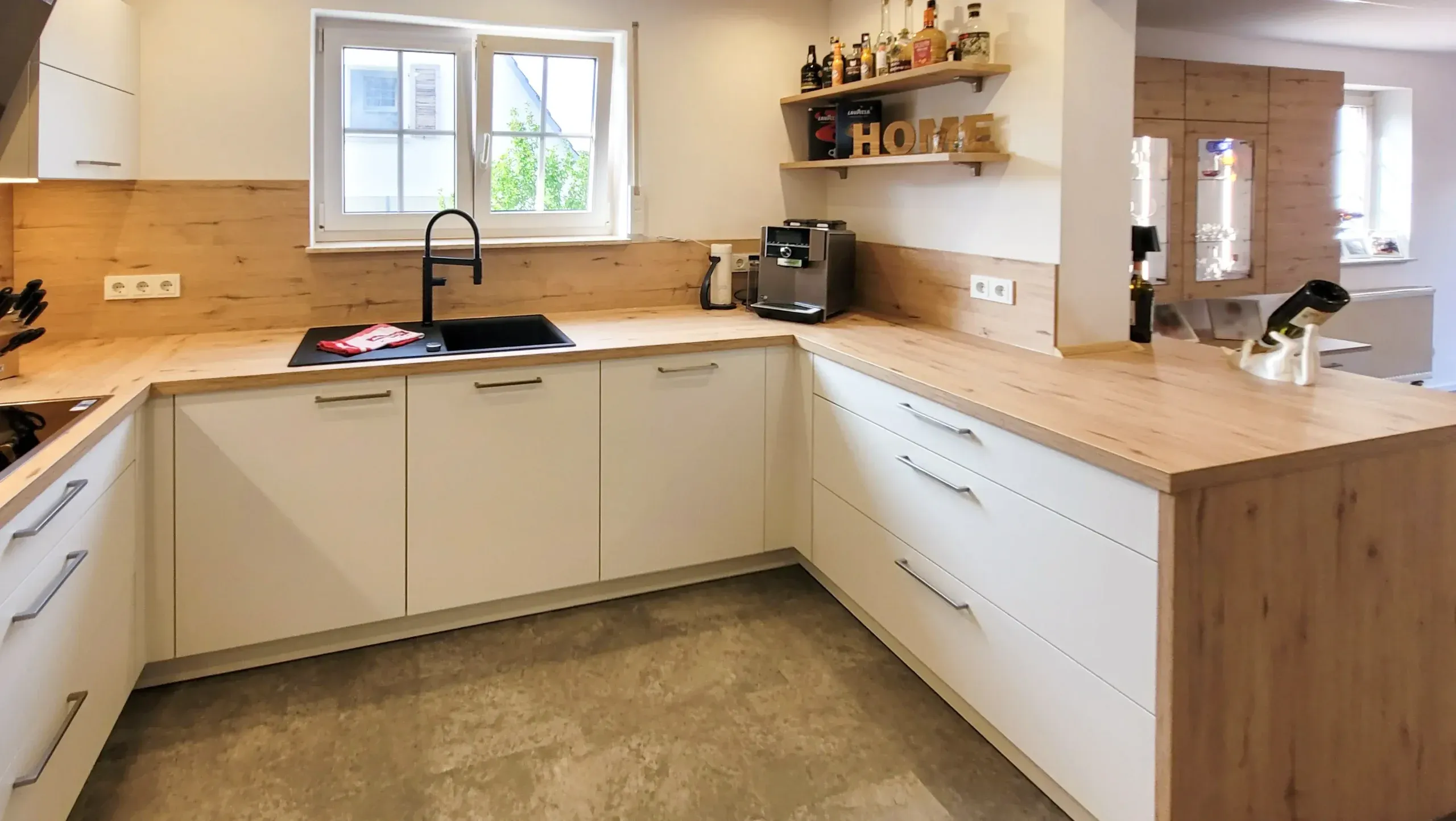 U-Küche mit Spülbereich am Fenster, schwarzer Doppelspüle und schwarzer Armatur, weißen grifflosen Unterschränken, Holz-Arbeitsplatte und Holz-Rückwand mit zwei Holzregalen