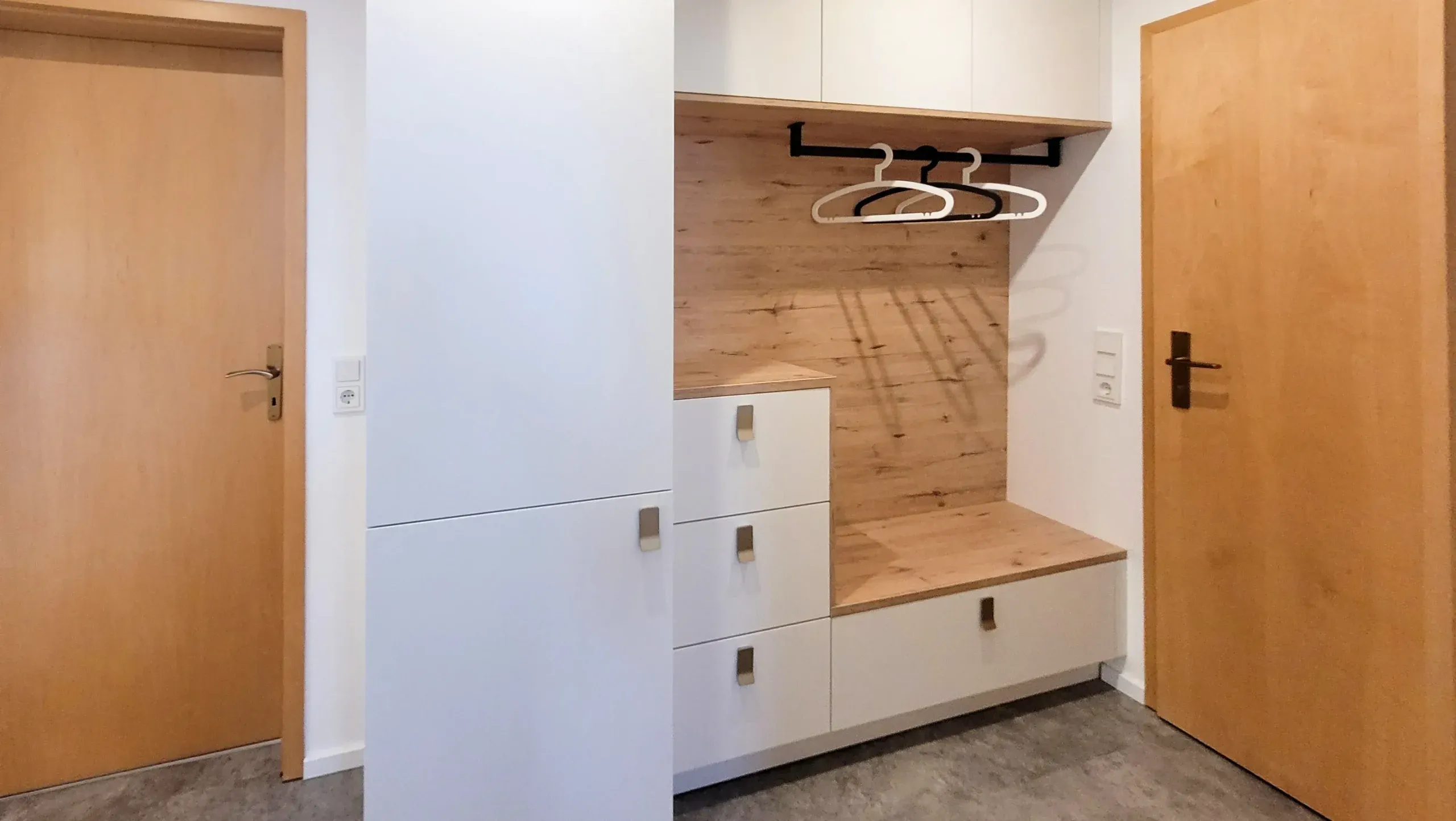 Garderobe im Eingangsbereich mit eingebauter weißer Schrankfront, Holz-Sitzbank mit offener Garderobennische und Schubladen, flankiert von zwei Holztüren