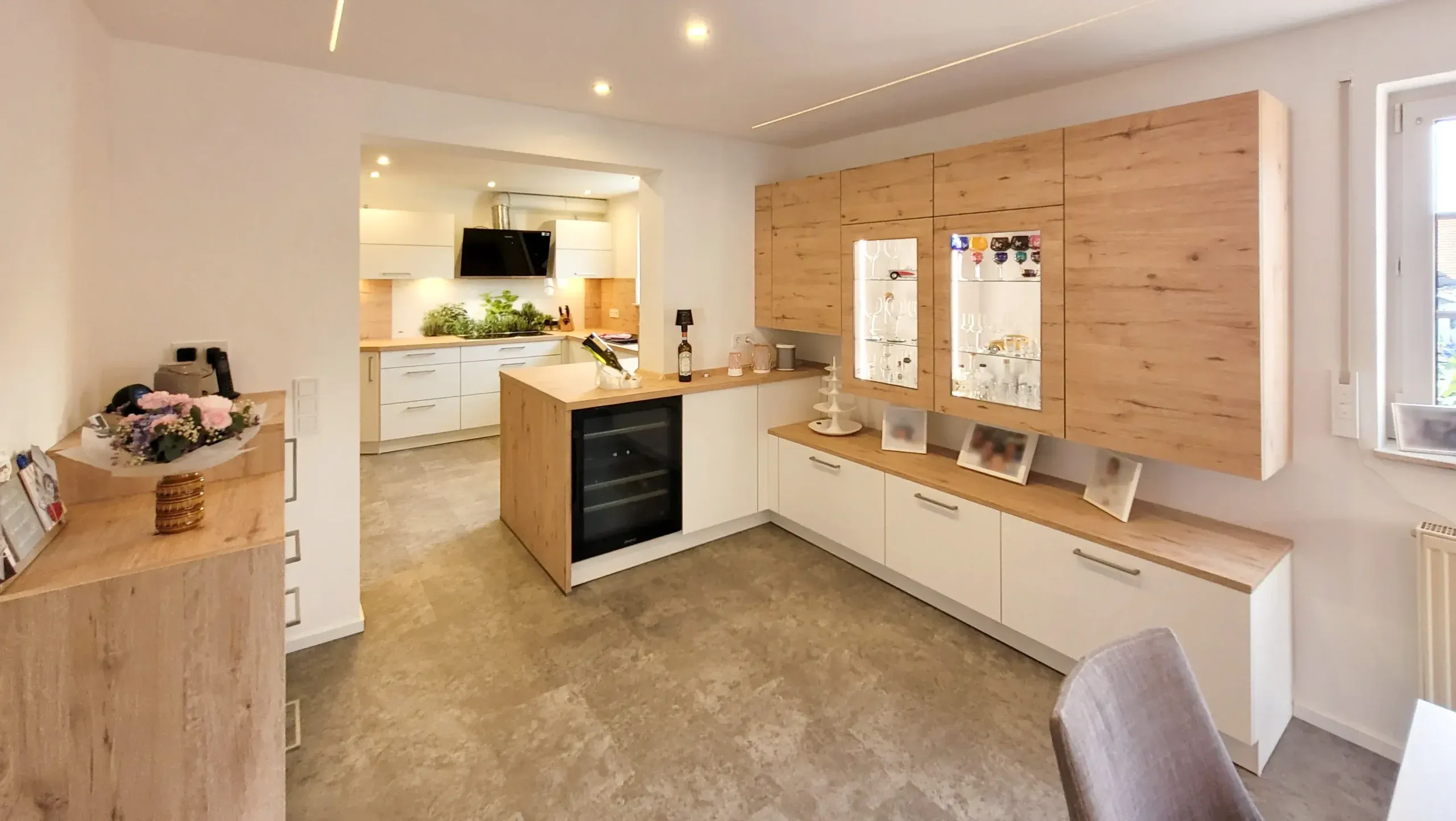 Wohnbereich mit langer Sideboard-Hochschrank-Wand aus Holz und Weiß, integrierten Glasvitrinen und Weinkühlschrank, im Hintergrund Blick in die Küche mit Insel und Wand-Dunstabzug