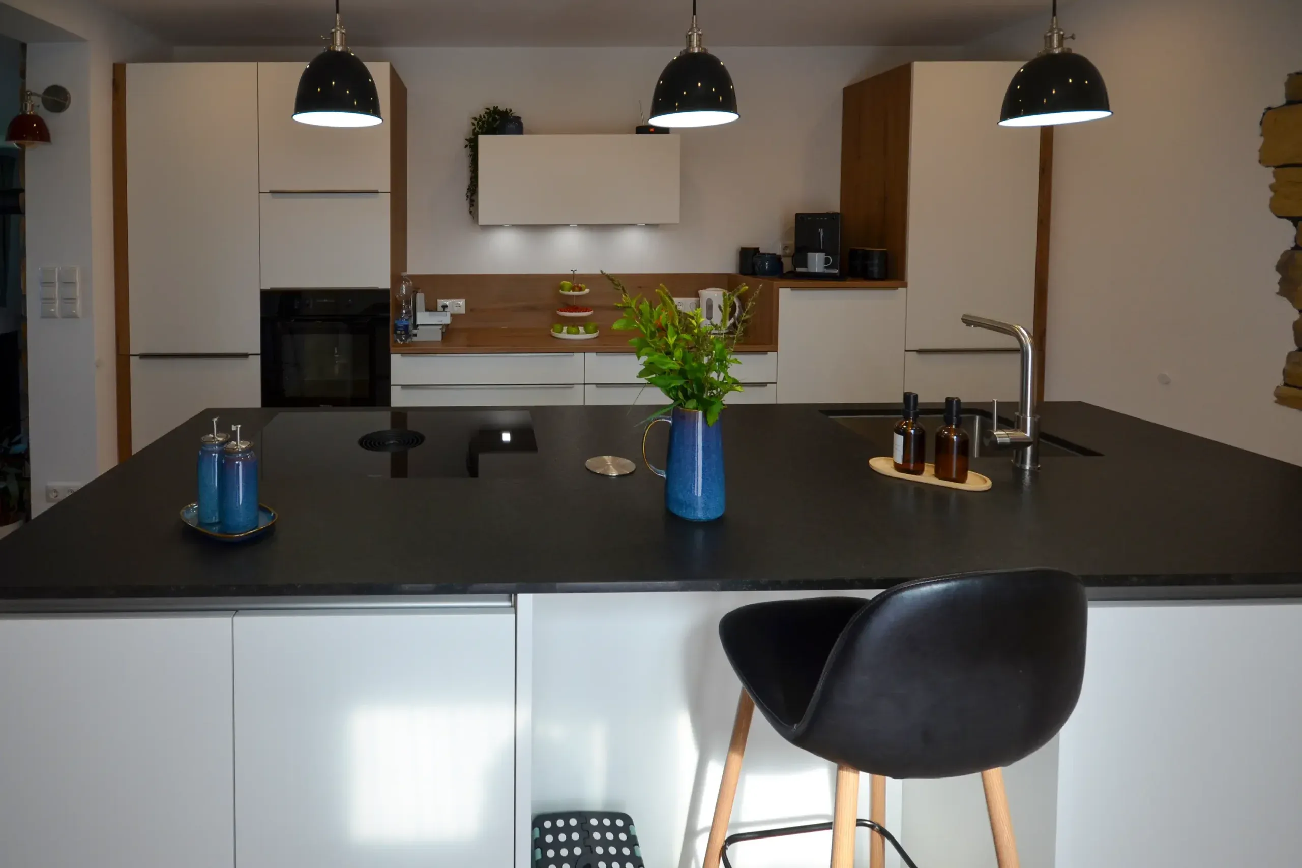 Detailansicht der Bartheke mit dunkler Arbeitsplatte und Barhocker im Vordergrund, blaue Deko-Vasen und Gewürzmühlen, dahinter unscharf weiße Schrankwand mit Backofen und Wildeiche-Element, drei schwarze Pendelleuchten – Referenzprojekt von Broßler Küche Aktiv von Dietmar Kraus