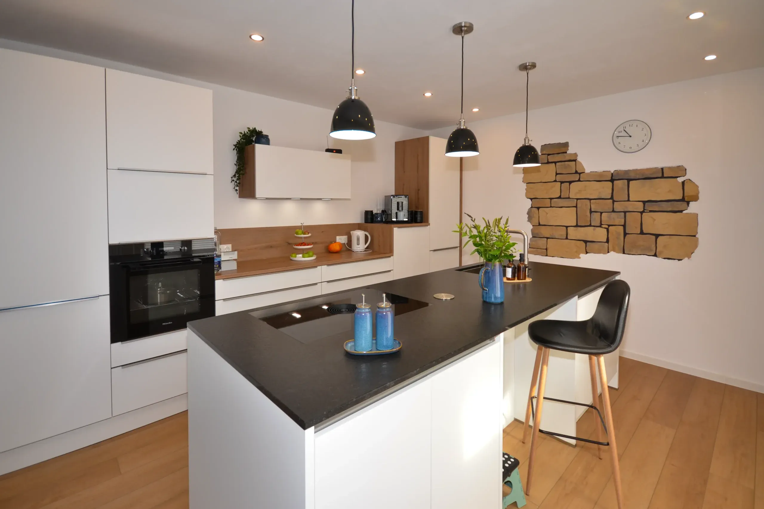 Moderne weiße Wohnküche mit Kochinsel mit dunkler Arbeitsplatte, Barhockern, Wildeiche-Nischenrückwand mit Backofen-Hochschrank, drei schwarzen Pendelleuchten und markanter Natursteinmauer mit Wanduhr im Hintergrund – Referenzprojekt von Broßler Küche Aktiv von Dietmar Kraus