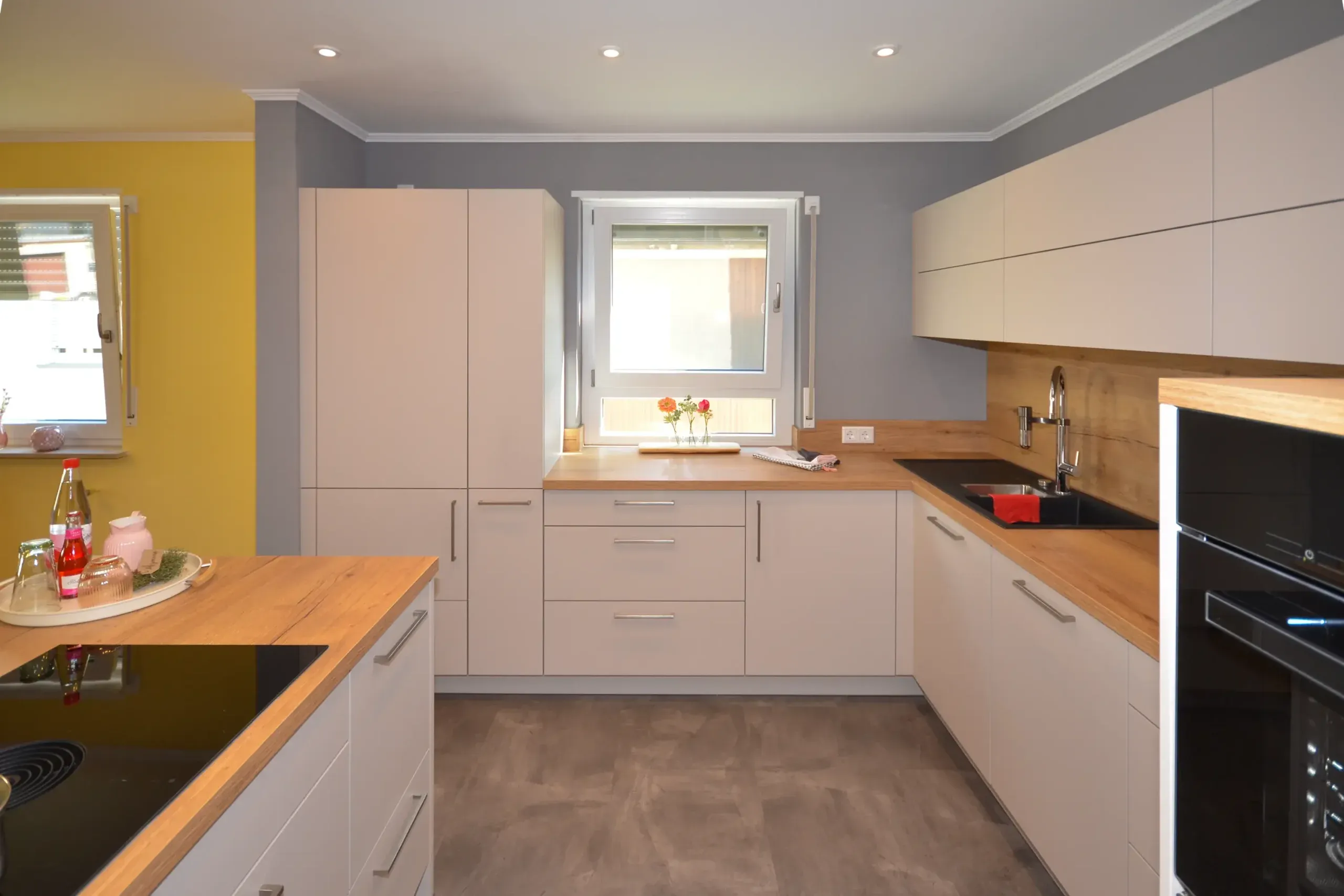 Moderne L-Küche mit beige/taupefarbenen matten Fronten, massiver Eichen-Arbeitsplatte, Spüle vor dem Fenster mit Blumendeko, integriertem Backofen rechts, Kochinsel links und gelber Akzentwand – Referenzprojekt von Broßler Küche Aktiv von Dietmar Kraus