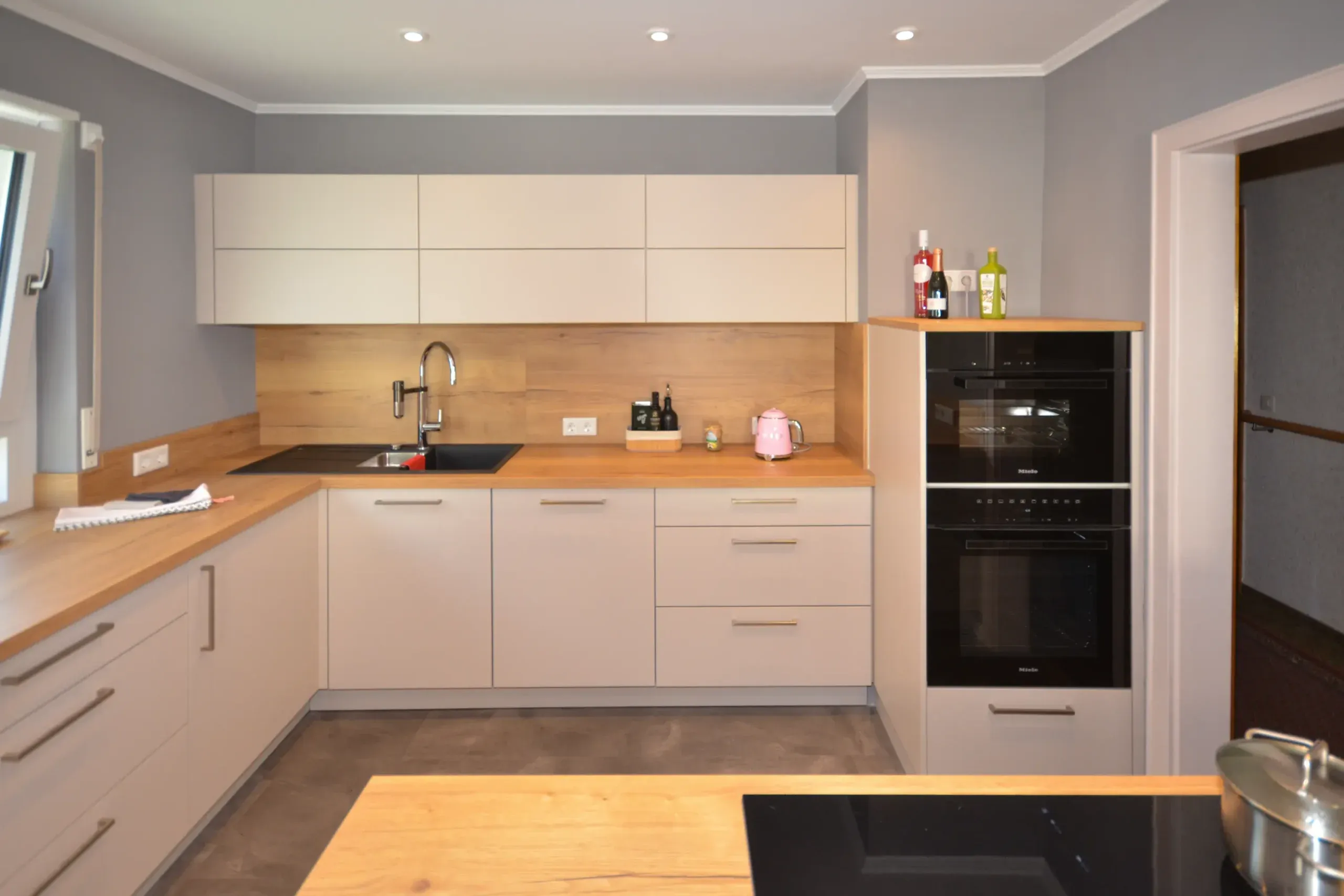Moderne Küche mit Spülenseite und Hängeschränken in beige/taupefarbenen matten Fronten, schwarzer Spüle in massiver Eichen-Arbeitsplatte, Holz-Nischenrückwand, Backofen-Hochschrank mit Backofen und Dampfgarer und Kochinsel mit Kochfeld im Vordergrund – Referenzprojekt von Broßler Küche Aktiv von Dietmar Kraus