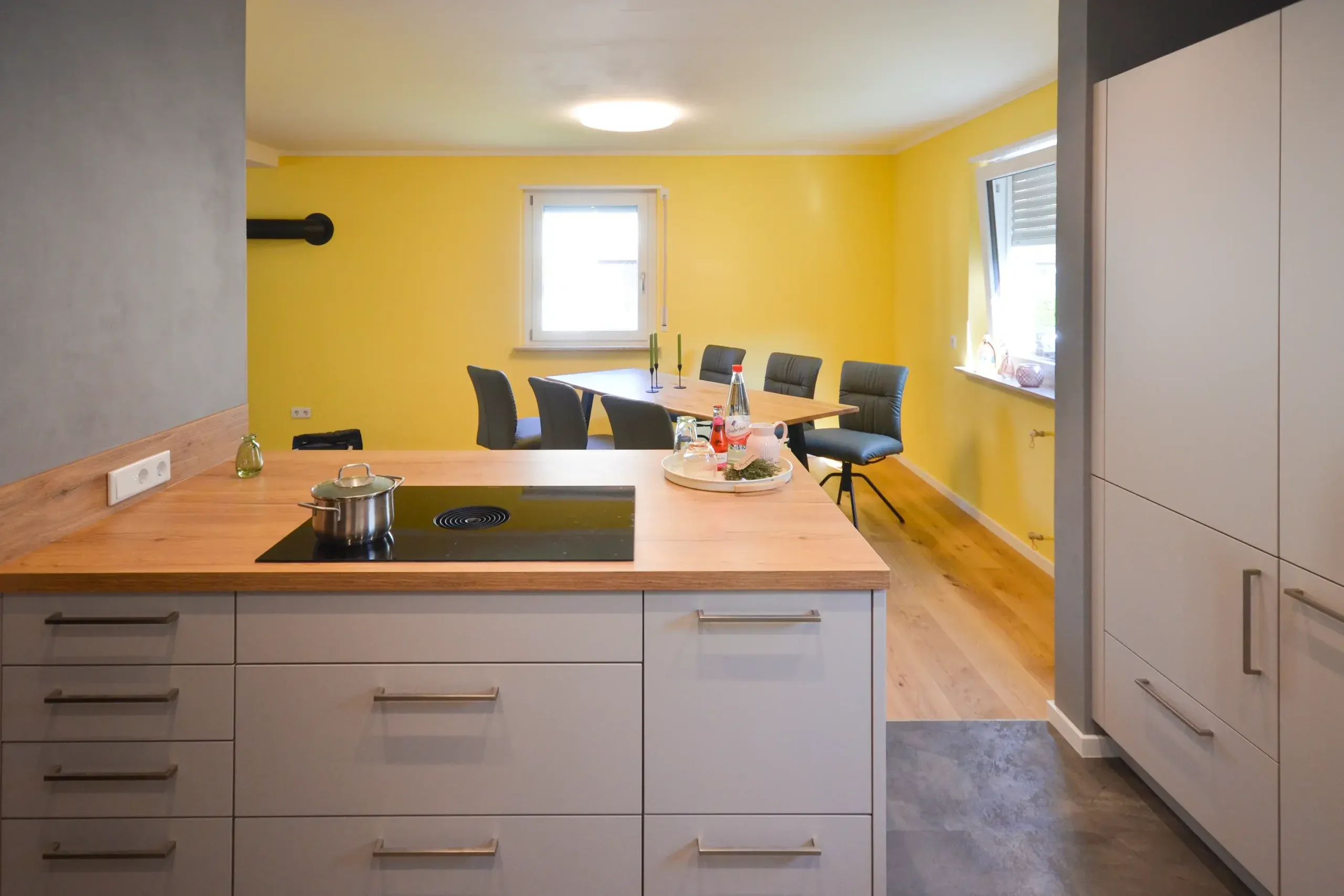 Moderne Wohnküche mit großer Kochinsel mit massiver Holz-Arbeitsplatte und Kochfeld, beige/taupefarbenen matten Fronten mit Bügelgriffen, dahinter Esstisch mit dunklen Stühlen vor gelber Akzentwand und Holzboden – Referenzprojekt von Broßler Küche Aktiv von Dietmar Kraus