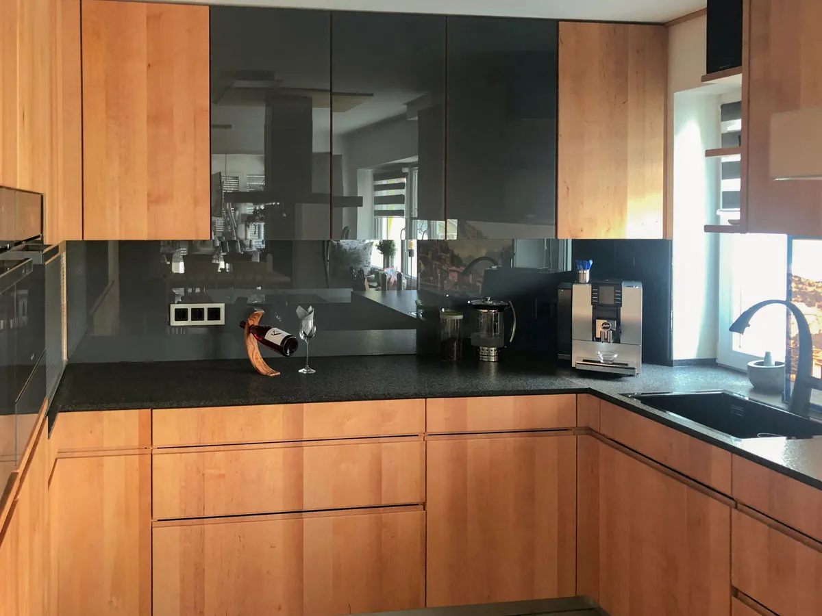 L-Küche in Erle-Holz mit Granitarbeitsplatte, dunkler Glas-Nischenrückwand und Spüle am Fenster