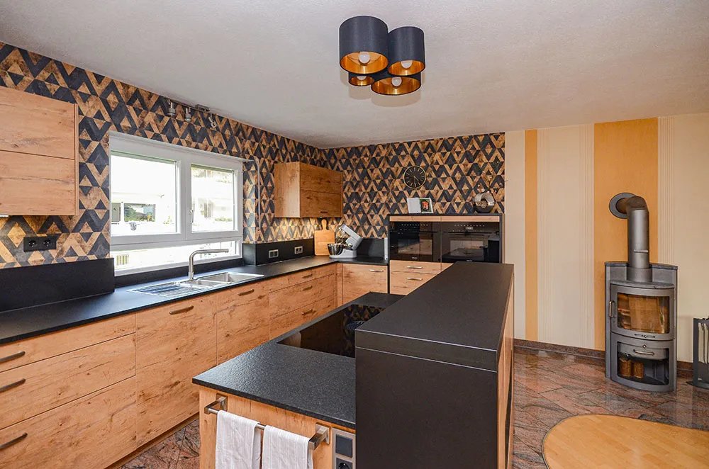 Küche in massiver Eiche-Altholz-Optik mit Kochinsel und schwarzer Steinarbeitsplatte, Arbeitszeile mit Spüle vor dem Fenster, blau-orange gemusterte Akzent-Tapete, dreifache Pendelleuchte mit Kupfer-Innenflächen, rechts Kaminofen im offenen Wohnbereich – Küchenplanung von Dietmar Kraus für Broßler Küche Aktiv
