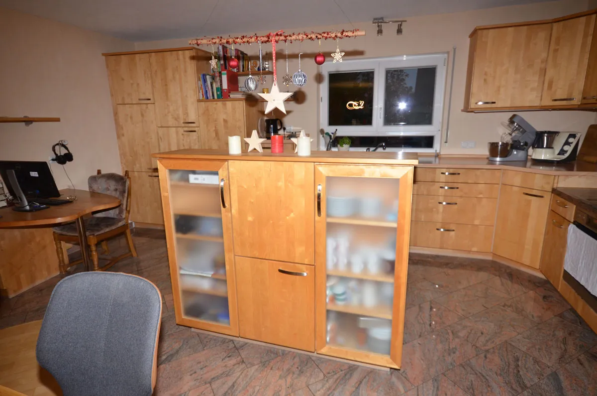 Vorher-Aufnahme einer Bestands-Küche in U-Form mit hellen Buchenholz-Fronten, dunkle Granit-Arbeitsplatte mit eingelassener Edelstahl-Spüle vor dem Fenster, Vitrinen-Sideboard mittig im Raum, gemusterter Steinboden, bewohnter Zustand mit weihnachtlicher Deko – Ausgangssituation vor der Küchenplanung von Dietmar Kraus bei Broßler Küche Aktiv