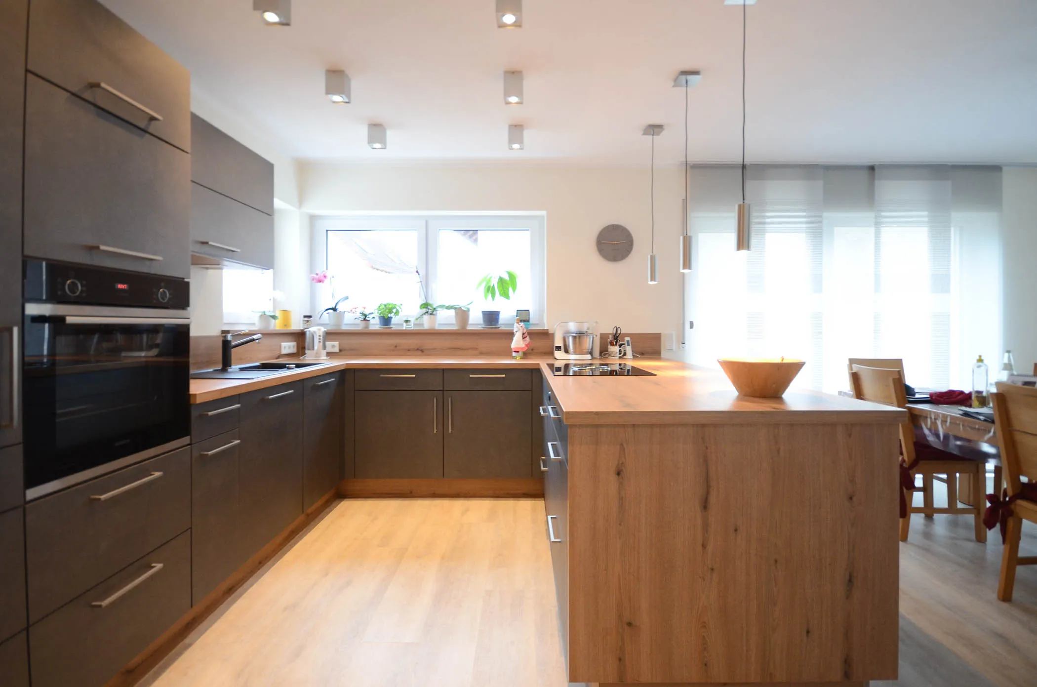 U-Küche mit dunklen Holzfronten und massiver Eichen-Arbeitsplatte als Halbinsel, integrierte Backofen-Hochschränke, Punktstrahler und Pendelleuchten, anschließender Essbereich – Referenzprojekt von Broßler Küche Aktiv