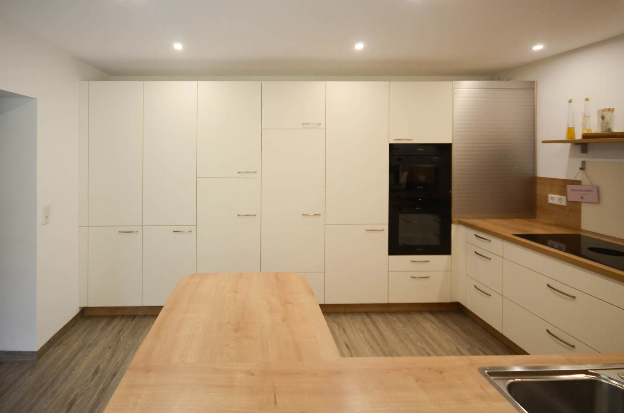 Weiße Küche mit raumhoher Hochschrankwand, integriertem Backofen, Halbinsel mit massiver Holz-Arbeitsplatte und Edelstahlspüle im Vordergrund – Referenzprojekt von Broßler Küche Aktiv