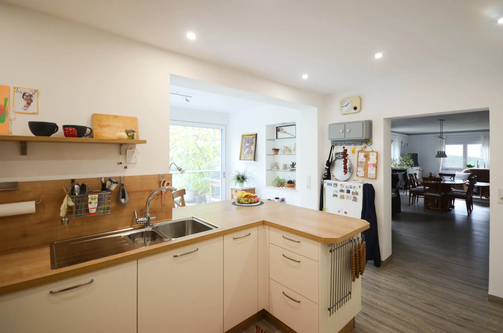 Weiße Wohnküche mit Doppelspüle, Holz-Arbeitsplatte, Holz-Nischenrückwand, Wandregal mit Deko und Durchgang zum Essbereich – Referenzprojekt von Broßler Küche Aktiv