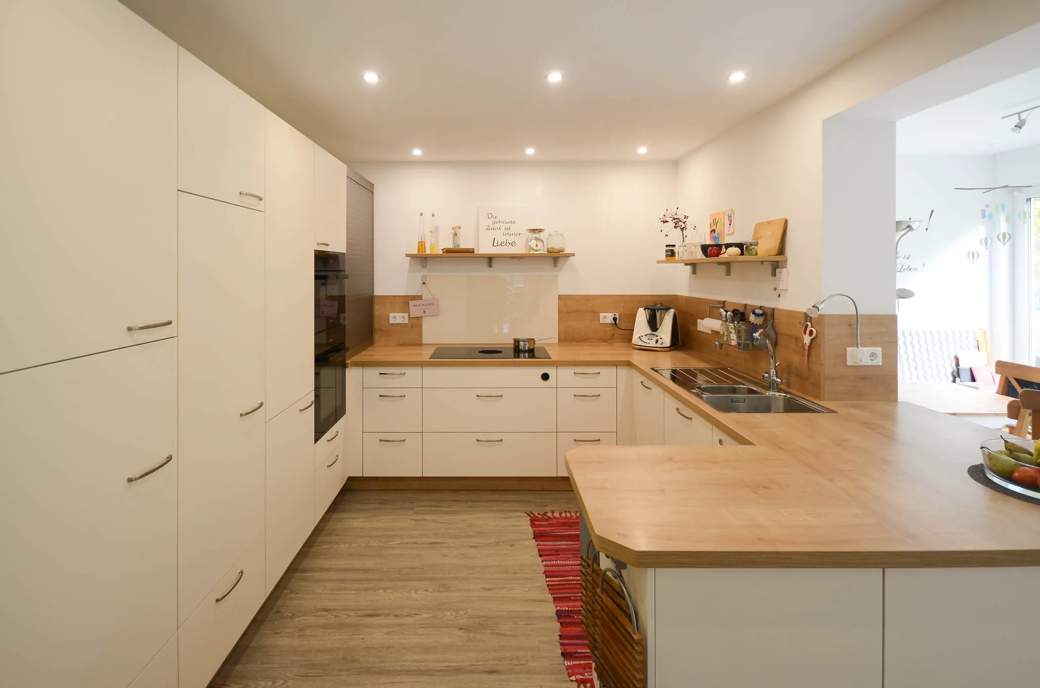 Großzügige weiße U-Küche mit massiver Holz-Arbeitsplatte, Spüle, Kochfeld in der Halbinsel, Holz-Nischenrückwand und kleinem Wandregal – Referenzprojekt von Broßler Küche Aktiv
